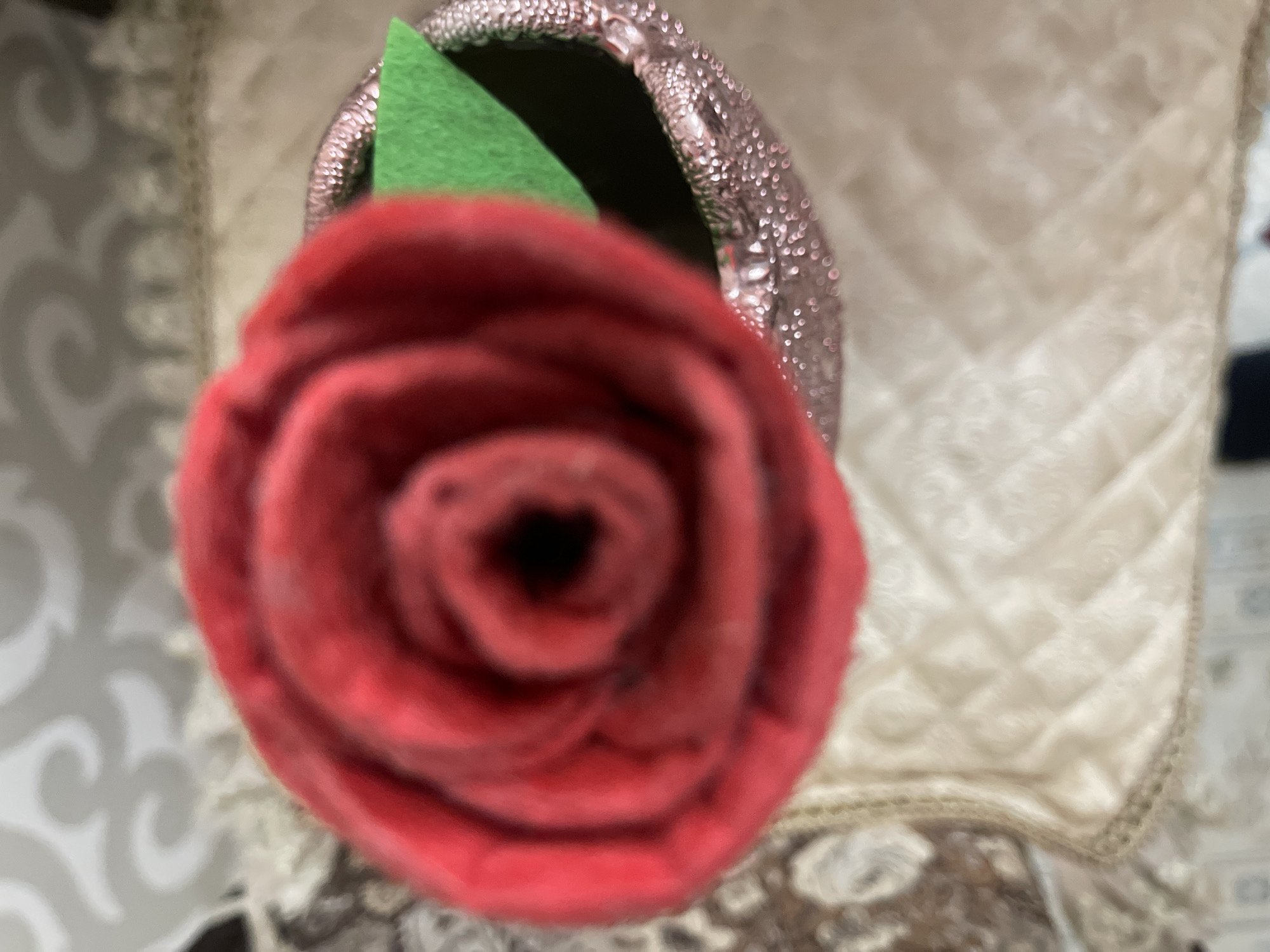 The image shows a close-up, top-down view of a red rose ornament in a glittery pink vase. A green felt leaf is visible emerging from the vase. The ornament and vase are placed on a textured, cream-colored fabric with a diamond pattern and a decorative border. In the background, a blurred pattern on a wall or fabric is visible, with hints of light and shadow. There are no people or discernible environmental features that would suggest a specific location within Bishkek City, Kyrgyzstan. The image is out of focus and does not provide enough detail to identify any specific landmarks or context for the city.