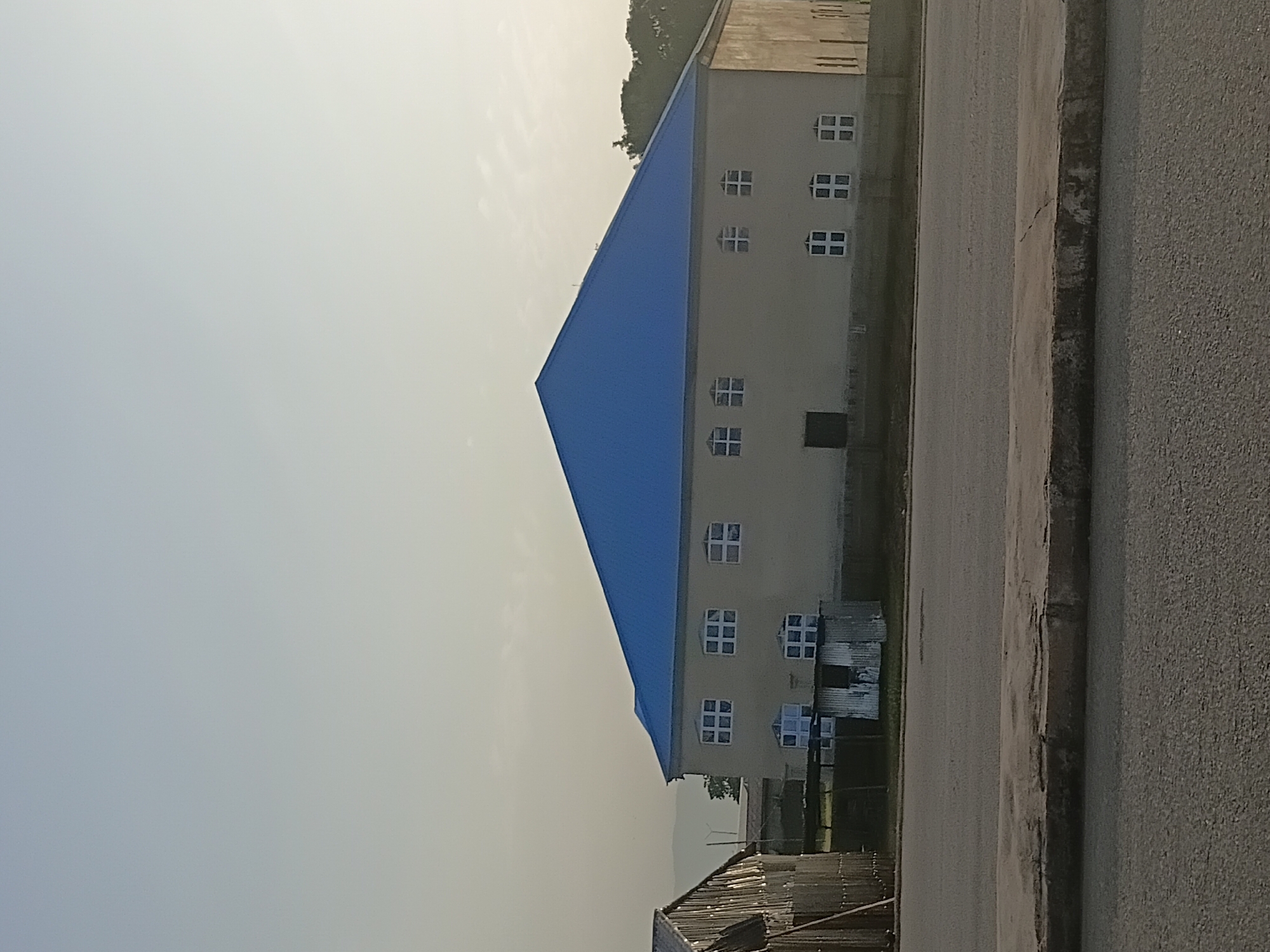 A light beige, two-story building with a bright blue, gabled roof stands in the background. The building has a dark rectangular door on the lower level and multiple windows on both floors, most of which are square and divided into smaller panes. The structure is situated next to a road and appears to be in a semi-urban or rural area, with some trees visible in the distance. To the left of the main building, a smaller, rustic structure with a corrugated metal roof is partially visible. The sky is clear and a light blue, indicating daytime. The overall scene suggests a quiet setting, possibly in the city of Jalingo, Nigeria, given the context of the prompt.