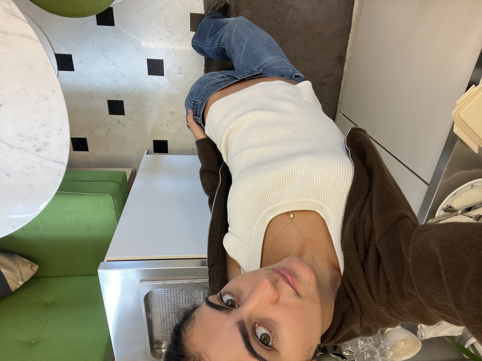 An inverted, high-angle selfie captures a young woman looking directly into the camera with a neutral expression. She is dressed in a white ribbed tank top, blue jeans, and a brown zip-up hoodie, and wears a delicate gold necklace with a small pendant. Her dark hair is pulled back.
The setting appears to be an indoor establishment, likely a cafe or restaurant, consistent with a Parisian context. To the left of the frame, a white marble tabletop is visible, alongside vibrant green cushioned seating, suggesting a booth or banquette area. The floor features a distinct black and white checkered pattern. To the right and behind the woman, a functional service area is visible, including a stainless steel counter with a built-in sink, white cabinetry or appliances, and various kitchen or serving items such as stacked plates, bowls, and clear glasses. The lighting in the space suggests it is daytime. There is no discernible text visible in the image.