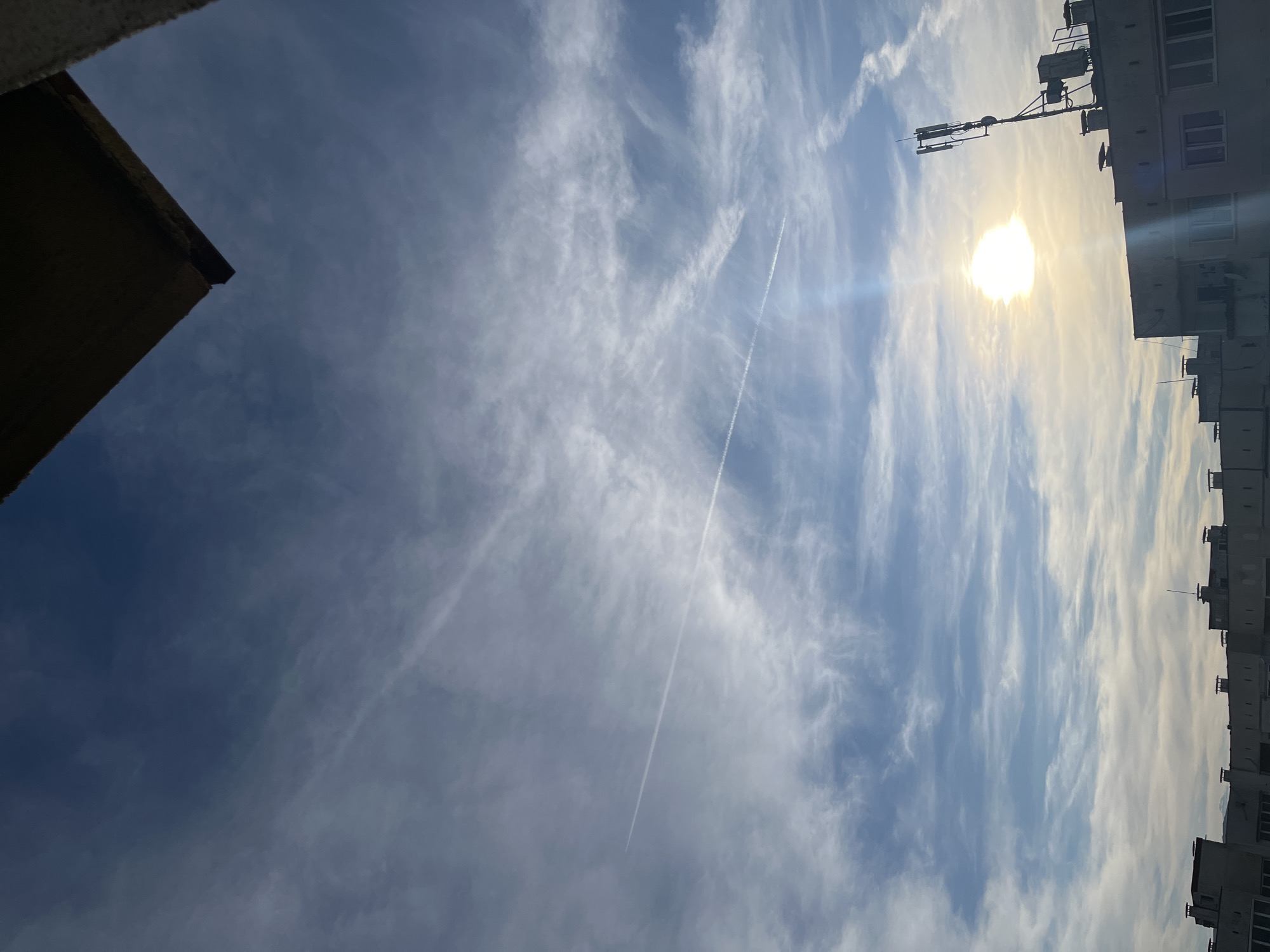 The image shows a low-angle view of the sky above an urban environment. The sky is predominantly blue with scattered wispy white clouds and a contrail from an airplane. The sun is visible, partially obscured by clouds, creating a bright glare. In the foreground, partial views of buildings are present. On the left, a section of a dark-colored building roof is visible. On the right, the side of a multistory building with several windows, balconies, and rooftop telecommunications equipment is visible. It can be identified as the city of Sofia, Bulgaria.