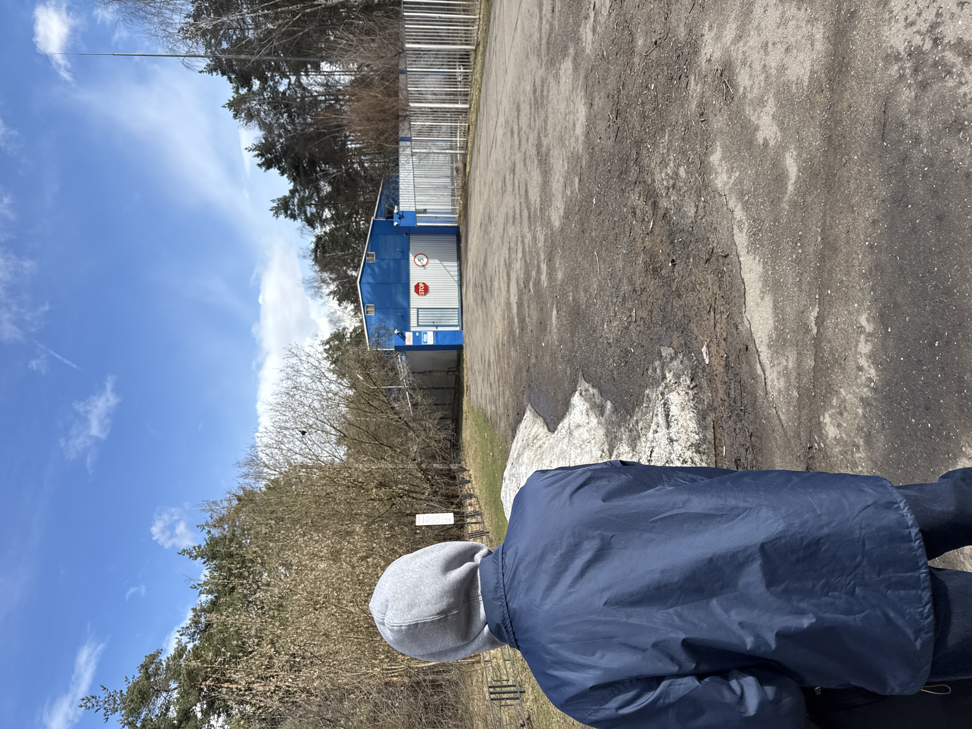 A ground-level photograph shows a person wearing a gray hooded sweatshirt under a navy-blue jacket, with their back to the viewer. The person is standing or walking on a paved area of asphalt.
In the background is a blue and white building with a stop sign visible. The building is adjacent to a high fence. Trees are visible on either side of the building and fence. Above the trees is a blue sky with scattered clouds.
The image suggests an outdoor setting, possibly a perimeter or access point in a built environment. There is no definite evidence to locate the image in Moscow, Russia.