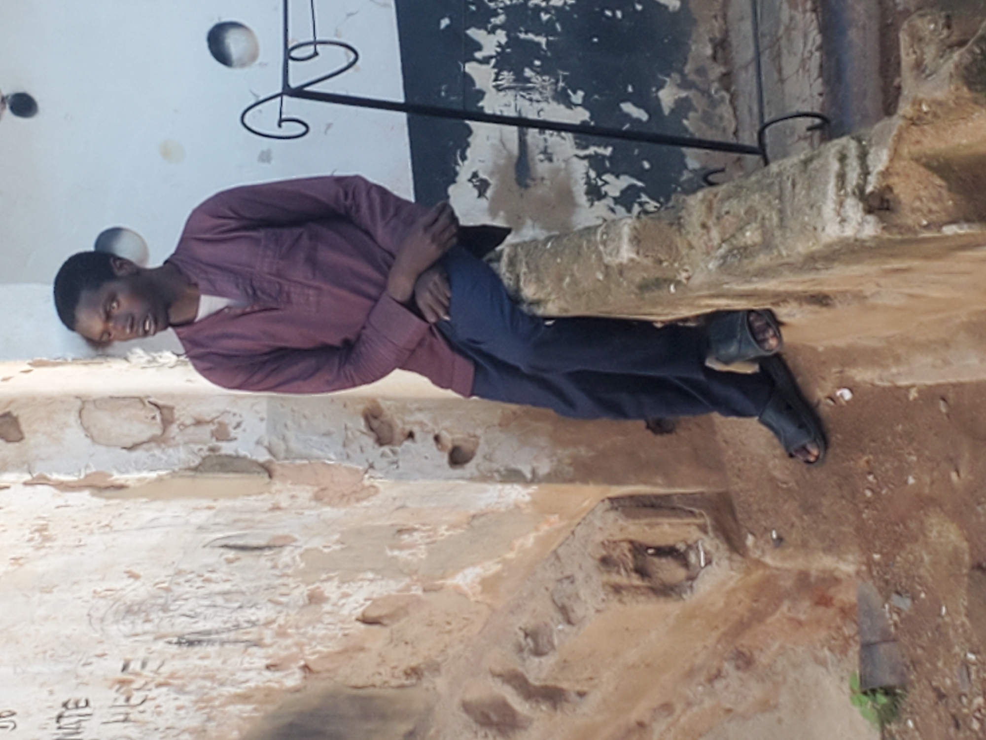 A full shot shows a young Black man sitting on a low concrete ledge outdoors in Gwong, Nigeria. The man is wearing a dark maroon button-down shirt with three buttons at the collar and dark blue pants. He has on black sandals. His arms are crossed and he is looking to his left and slightly upward. The wall behind him is peeling and discolored with what appears to be graffiti. To his right, a black metal railing with a decorative scroll pattern curves upward from the ground. The ground is covered with dirt and small rocks.