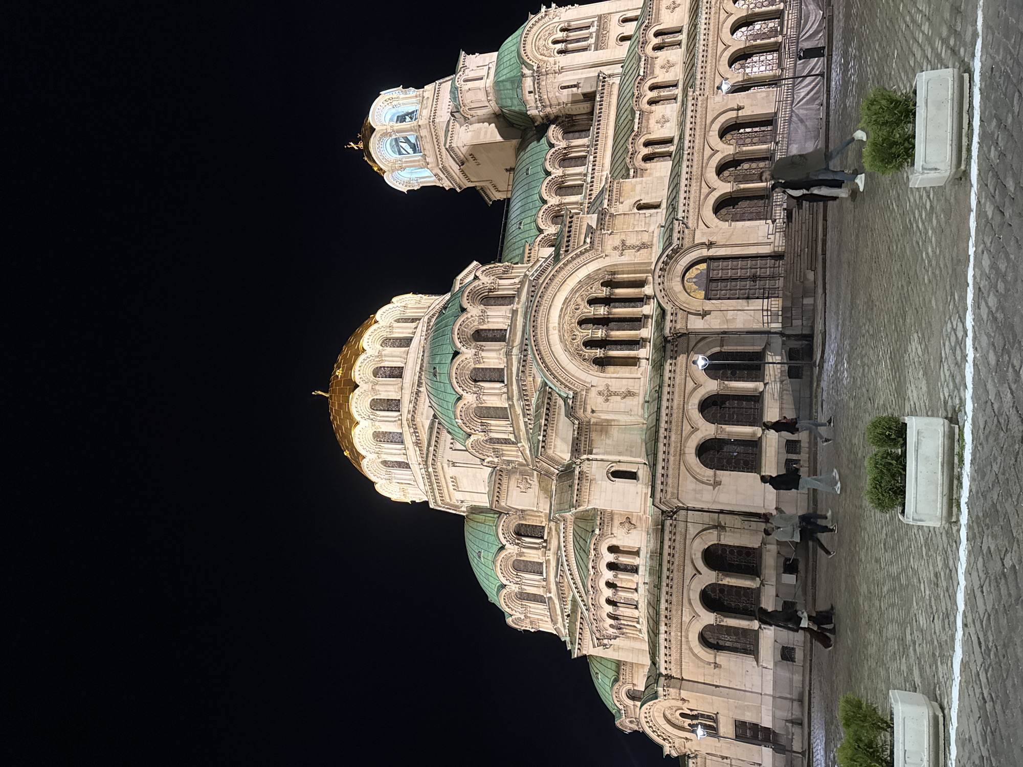 The image depicts the Alexander Nevsky Cathedral in Sofia, Bulgaria at night. The cathedral is illuminated, revealing its intricate architectural details, including its domes, arches, and windows. Several people are walking along the cobblestone pavement in front of the building. There are rectangular planters filled with greenery lining the pavement. The background is mostly black, indicating the nighttime setting.