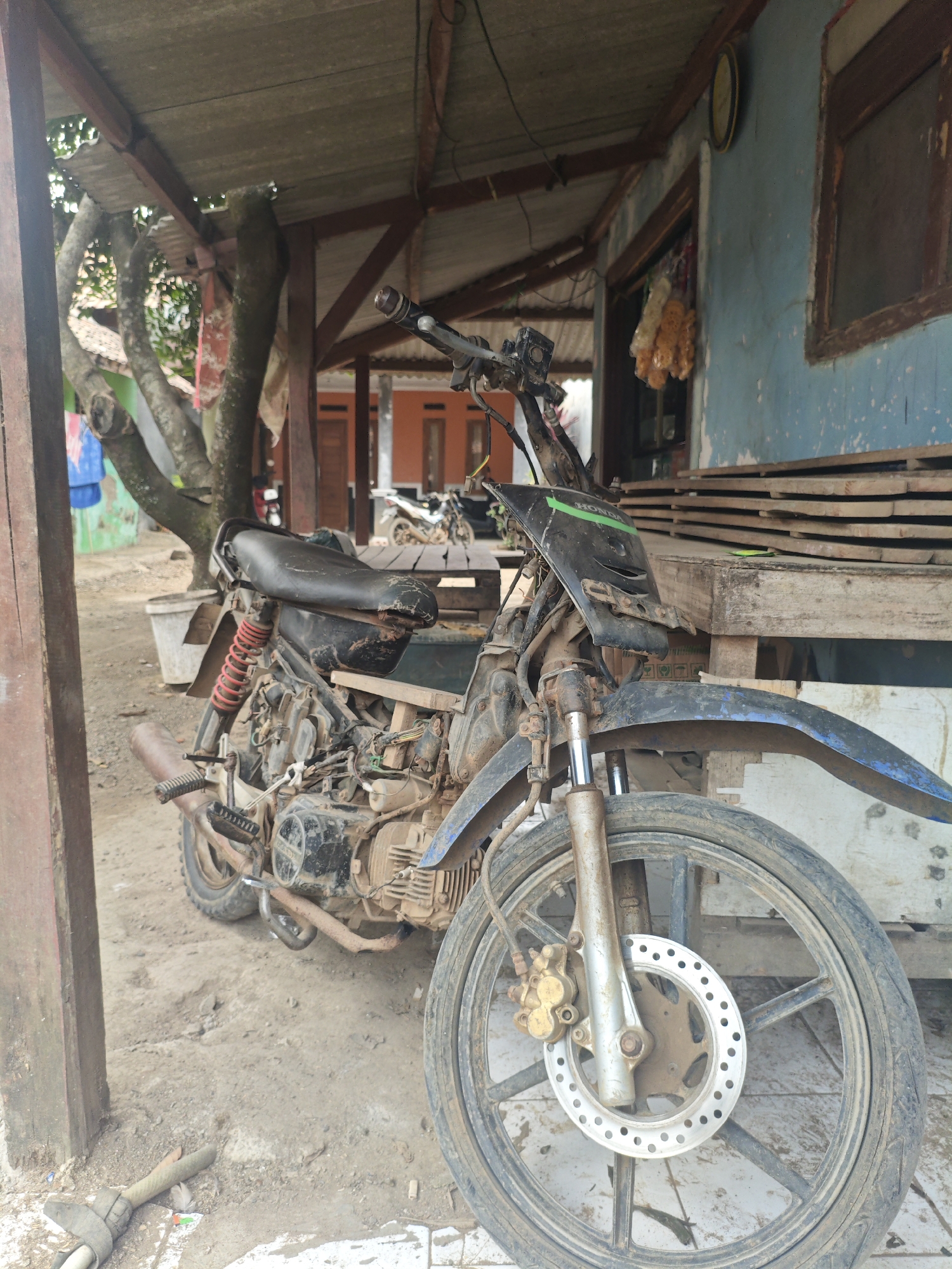 The image depicts a weathered, black motorcycle parked under a rustic, open-air shelter. The motorcycle appears dirty and somewhat disused, with exposed engine parts and a worn seat. Red shock absorbers are visible on the rear suspension. The shelter's roof is corrugated metal, supported by wooden beams, and the ground is dirt and some concrete. To the right of the motorcycle, a light blue wall with a wooden window frame is visible. Some wooden planks are stacked against the wall. In the background, through an opening in the shelter, another motorcycle and the facade of a house with orange walls and doors can be seen. The overall setting suggests a rural or semi-rural environment, possibly a yard or roadside in Bantarwaru, Indonesia, given the architectural style and tropical vegetation hinted at by the trees. No people are present in the image.
