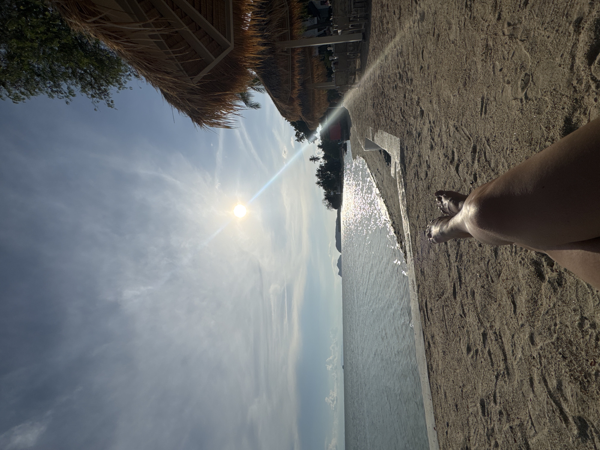 The image shows a seaside scene in Baan Tai, Thailand, viewed from a low angle. The foreground features sand and a partial view of a person's leg and feet. The middle ground consists of the sea, reflecting sunlight, bordered by a concrete structure. In the background, trees and beachside structures like thatched umbrellas are visible. Above, the sky is blue with scattered clouds and the sun emitting a bright glare.
