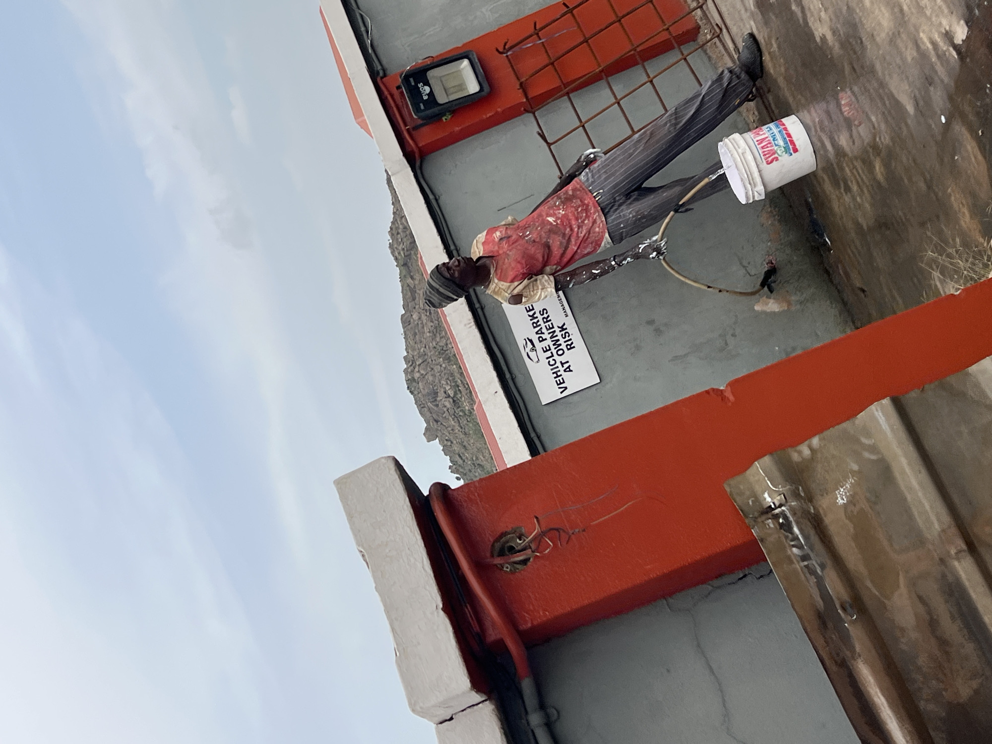 The image, likely taken in Gura Top, Nigeria, presents a low-angle view of a structure. A scarecrow-like figure stands near the edge of what appears to be a building. The figure is dressed in striped trousers, a red shirt, and a hat, holding a Swan brand bucket from which water is flowing. A sign is visible with the text "VEHICLE PARKE AT OWNERS RISK". The structure has red and grey paint. Wiring is exposed in one area. The background includes a cloudy sky.