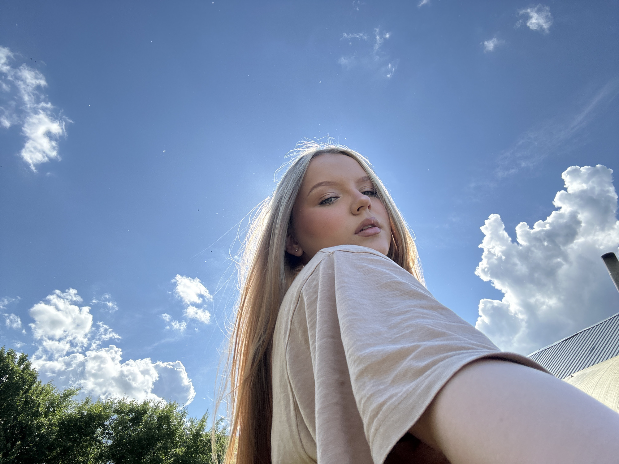 A low-angle, outdoor photograph shows a blonde woman in a beige shirt, looking over her shoulder at the camera. The backdrop is a bright blue sky dotted with scattered cumulus clouds and sunlight. The lower left corner of the frame contains green foliage, suggesting trees or shrubs. In the lower right, a structure with a silver corrugated roof and a chimney-like extension is partially visible. Due to the presence of the roof, foliage, and cloudy sky, and based on the presence of buildings in Chaikovsky, Russia, this image was possibly taken on the outskirts of the city or near a park.