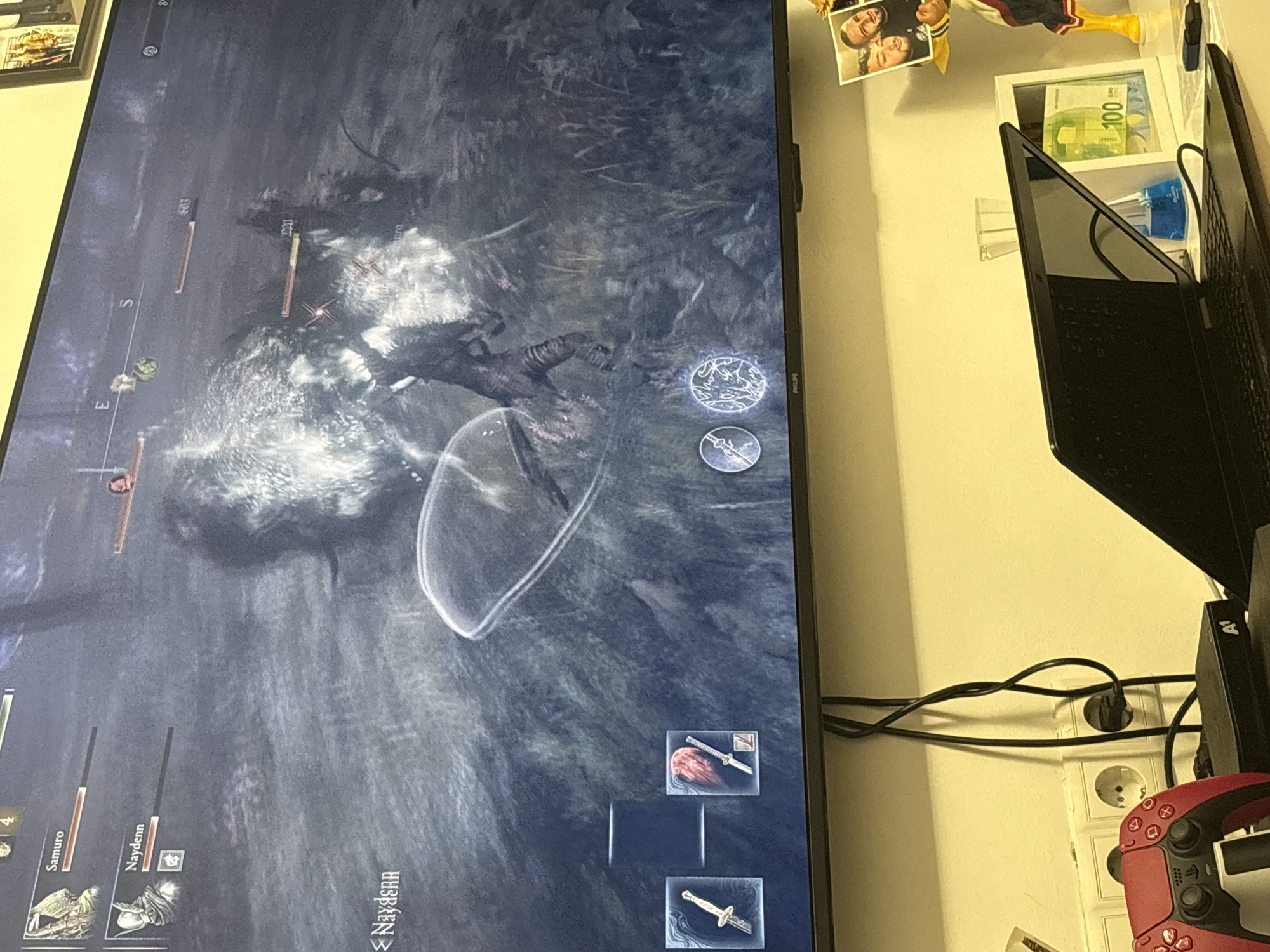 The image shows a television screen displaying a video game. The game appears to be set in a dark, fantasy environment, and a character is engaged in combat. The screen is bordered by a narrow bezel and is positioned on a wall. To the right of the television are several items, including a laptop, a picture frame with a banknote inside, a picture of two people, a PlayStation 5 controller, a power outlet strip with multiple plugs, and various wires. The location in the city of Plovdiv, Bulgaria, cannot be determined from the image.