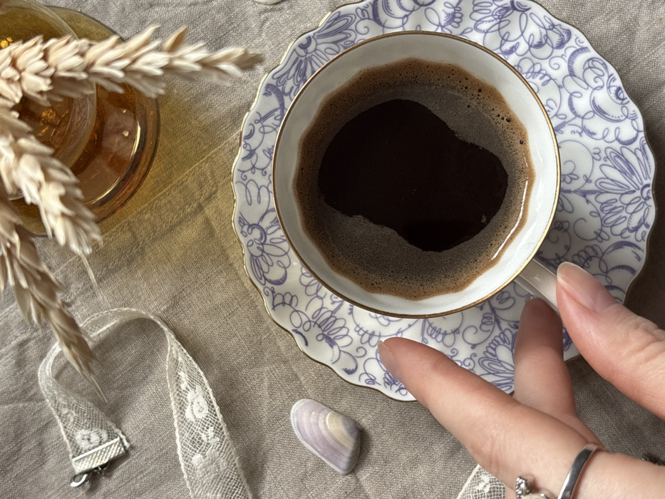 An overhead view depicts a domestic scene in Kudrovo, Russia. A white ceramic coffee cup with a scalloped gold-rimmed edge contains dark coffee topped with a layer of light brown foam. The cup rests on a matching white saucer adorned with a blue floral and swirl pattern, also featuring a gold rim. A human hand, positioned from the lower left, grips the cup's handle with its thumb and index finger; a silver ring with a white spherical gem is visible on the ring finger. The arrangement is situated on a light beige, woven linen-textured surface. To the upper left, a piece of cream-colored lace ribbon with a silver clasp lies partially unfurled. Below the ribbon, an elongated, polished stone exhibiting faint purple and white striations rests on the fabric. In the upper right quadrant, a translucent amber-colored glass vase containing several dried, light brown wheat stalks is partially visible.