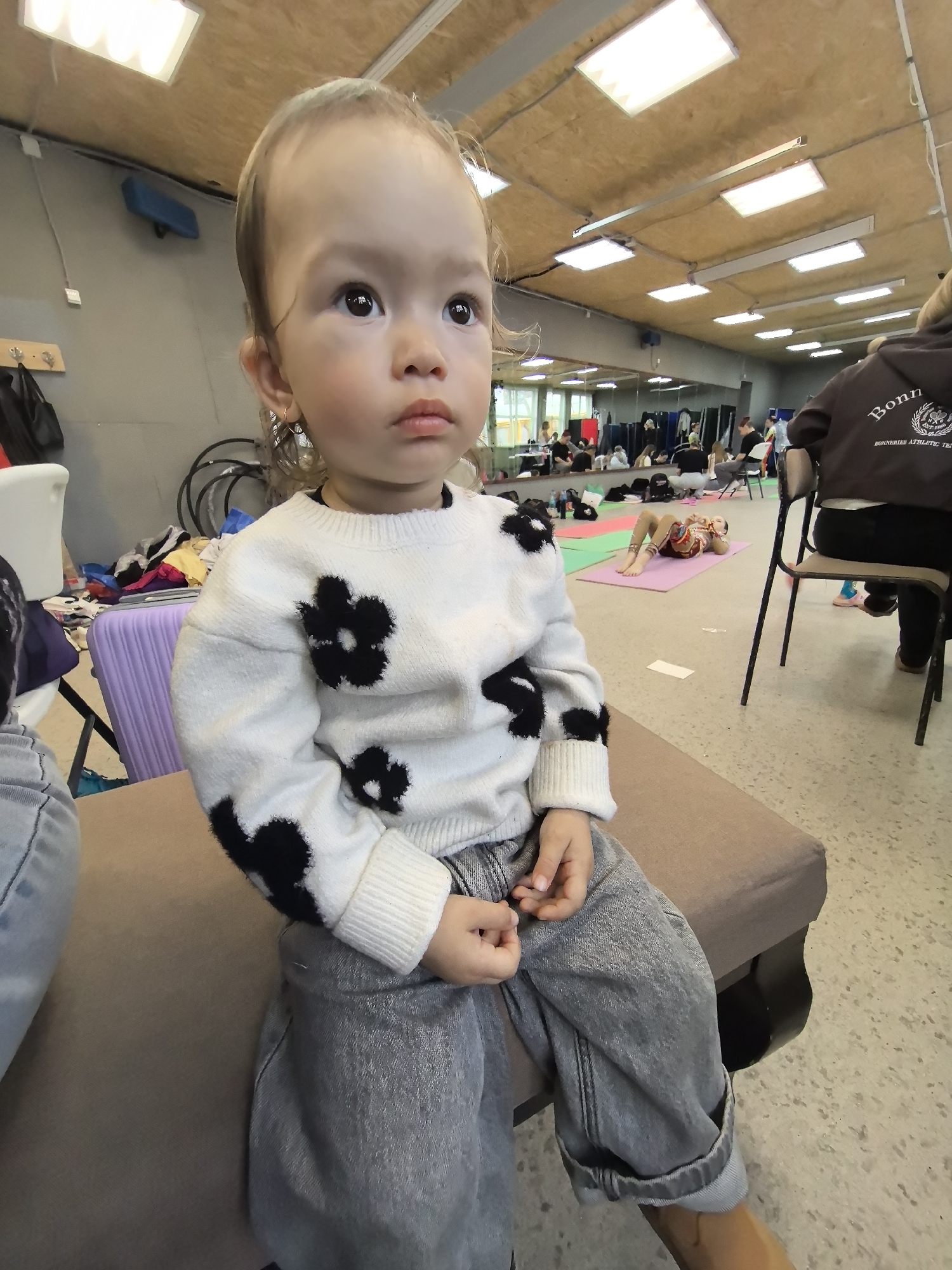 A young child wearing a white sweater with black flower designs and grey jeans sits on a brown upholstered bench. The location appears to be a gymnasium or dance studio, given the mirrored wall, exercise mats, and people stretching in the background. One person is lying on a purple yoga mat. The lighting is bright and artificial. A person wearing a hoodie labeled "Bonn" is sitting in the background. There is a suitcase next to the child. The child appears to be looking off to the side. The location is not in Izhevsk, Russia.