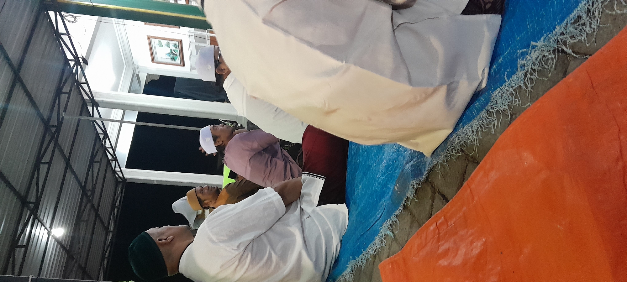 The image depicts an outdoor gathering, likely at night given the artificial lighting and dark background. Several men are seated on blue and orange mats, facing away from the camera. They appear to be engaged in a religious or cultural event. The men are wearing traditional Islamic attire, including kufis of various colors and long, loose-fitting garments. One man in the foreground is wearing a green kufi and a white shirt. Behind him, another man wears a brown shirt and a tan kufi. Further back, a man in a purple shirt and white kufi is visible, as is a man in a white shirt and white kufi. The environment suggests a covered outdoor space, with a metal roof supported by white pillars. In the background, a framed picture hangs on a white wall. The location is identified as Bendungan, Indonesia.