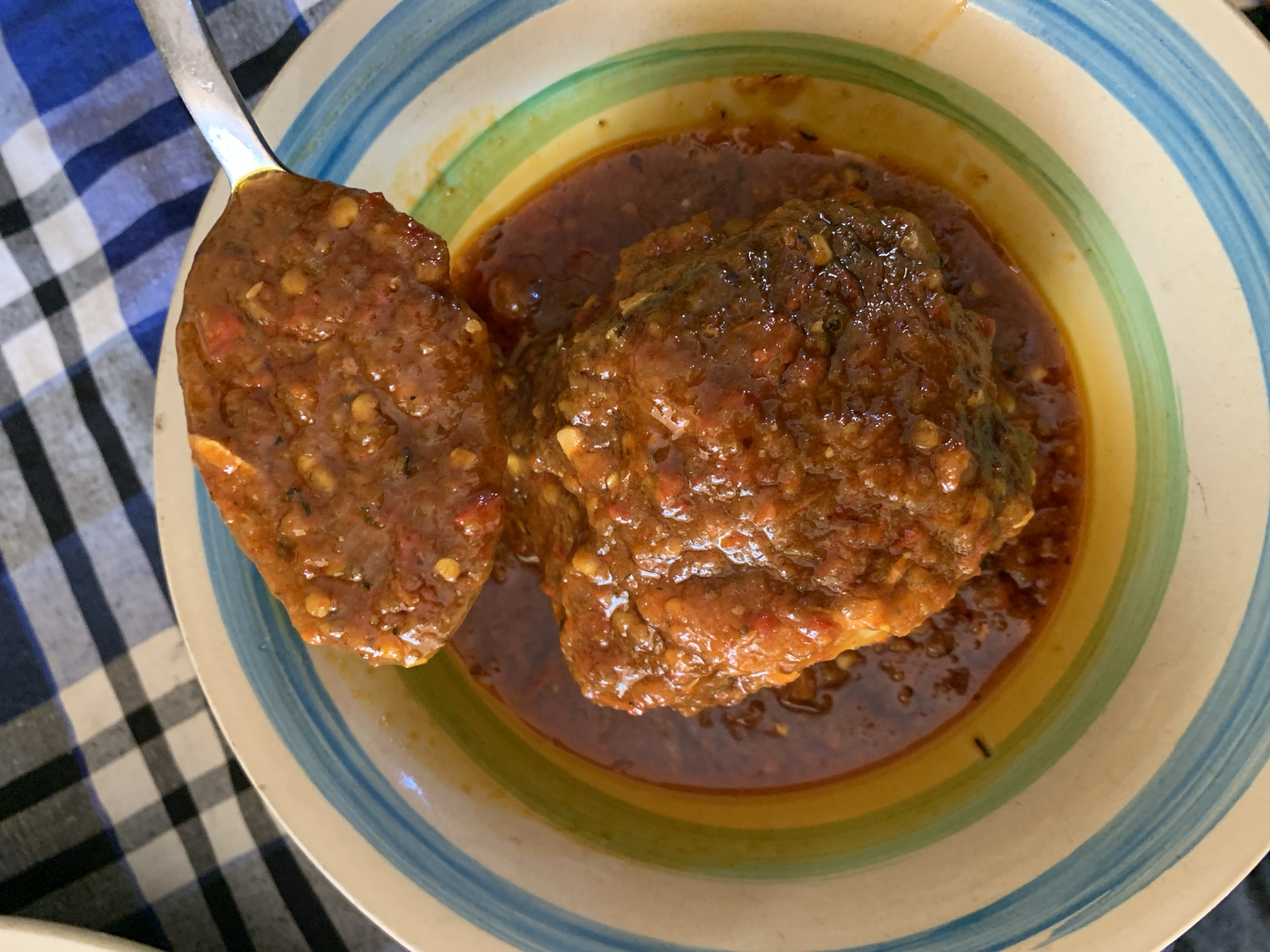 The image displays a close-up, overhead view of a hearty meal served in a colorful ceramic bowl, situated in the city of New Stadium, Nigeria. The primary subject is a reddish-brown, chunky stew or sauce, likely containing meat or fish, which is largely submerged within its own rich gravy. Visible within the stew are numerous small, irregular flecks of a lighter red or orange color, suggestive of chili flakes or other spices. A silver-colored metal spoon is positioned above the bowl, holding a substantial portion of the same thick, reddish stew, lifted slightly as if in the process of serving or eating. The ceramic bowl features a cream-colored base with a distinct rim decorated with horizontal stripes in shades of blue, light blue, green, and yellow. In the upper left background, a portion of a checkered tablecloth is visible, characterized by alternating squares of dark blue or black and white. No individuals are depicted in the scene.