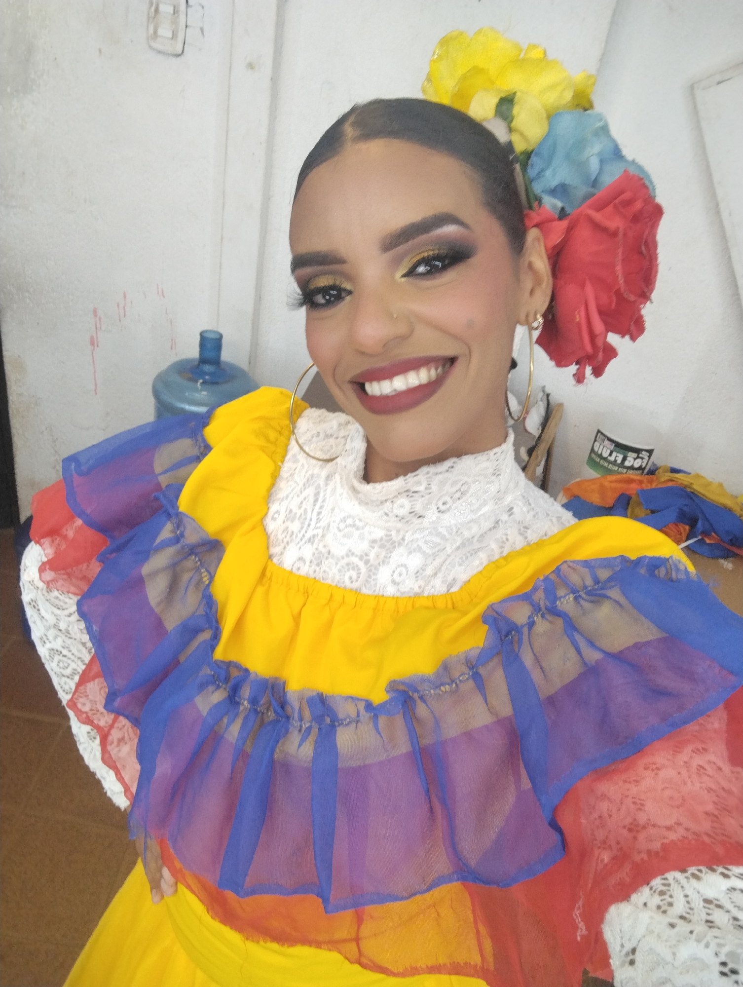 A woman is smiling directly at the camera, dressed in a vibrant, traditional Venezuelan costume. The dress features a yellow bodice and skirt, a white lace high-necked collar, and a multi-layered ruffled yoke in red, blue, and yellow. She has large gold hoop earrings and flowers in her hair. Her makeup is prominent, with heavily defined eyebrows, gold eyeshadow, and dark red lipstick. In the background is a water cooler, a wall, and some fabric that appears to match the dress. The setting appears to be indoors.