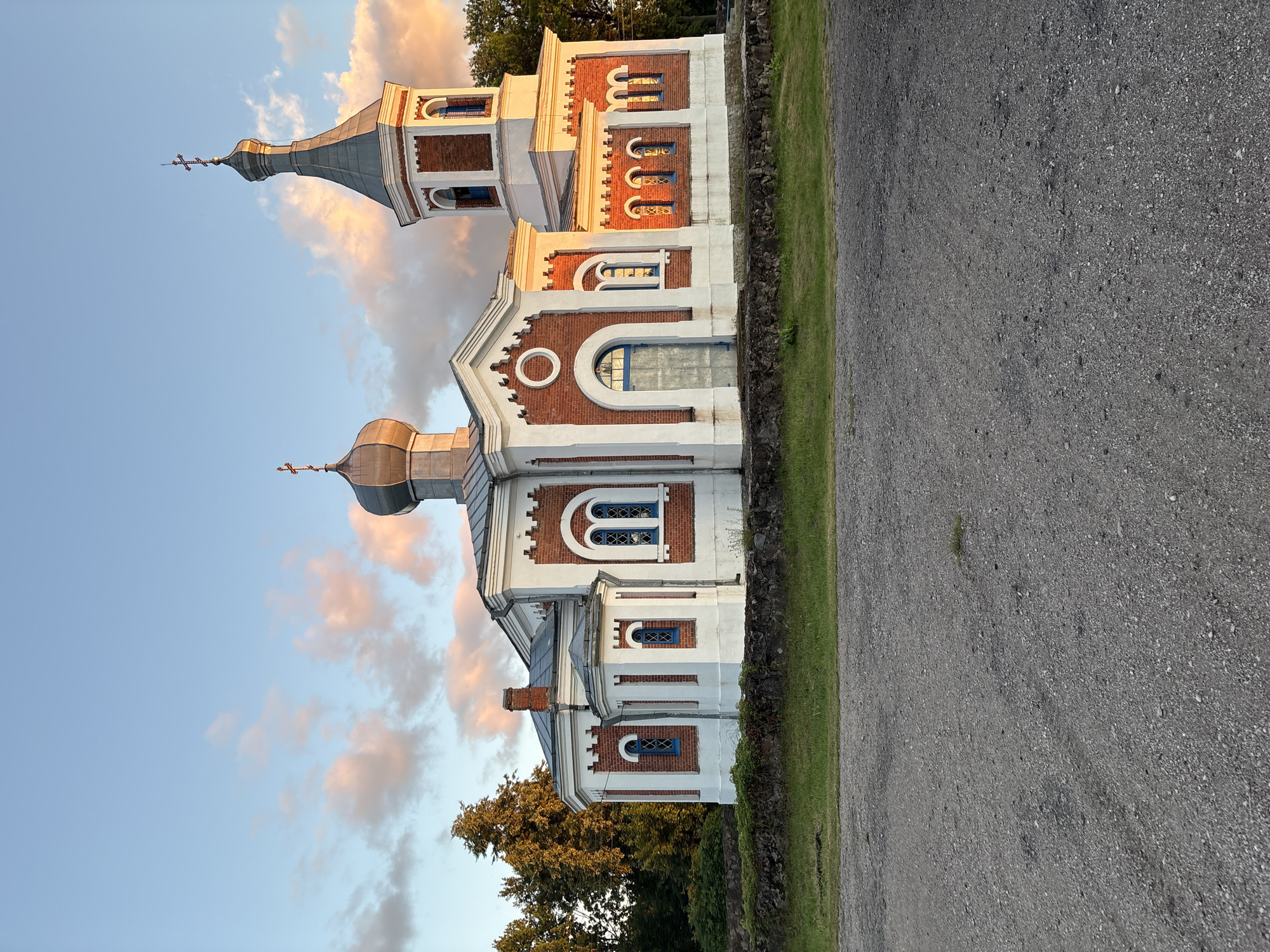 The image depicts an Eastern Orthodox church with white and red brick architecture, situated in the village of Krupets, within the Vidza rural council, Braslaw District, Vitebsk Region, Belarus. The church features multiple domes, including a prominent grey dome and a smaller copper-colored dome, both topped with crosses. A bell tower, also with a cross-topped spire, is attached to the right side of the building. The structure is adorned with blue-trimmed arched windows and decorative brickwork. In front of the church, a low stone wall separates it from a grassy area, and beyond that, a gravel road occupies the foreground. The sky above is a pale blue with scattered, softly lit clouds in hues of pink and orange, suggesting it is either sunrise or sunset. Lush green trees are visible in the background, to the left and right of the church. No people are present in the image.