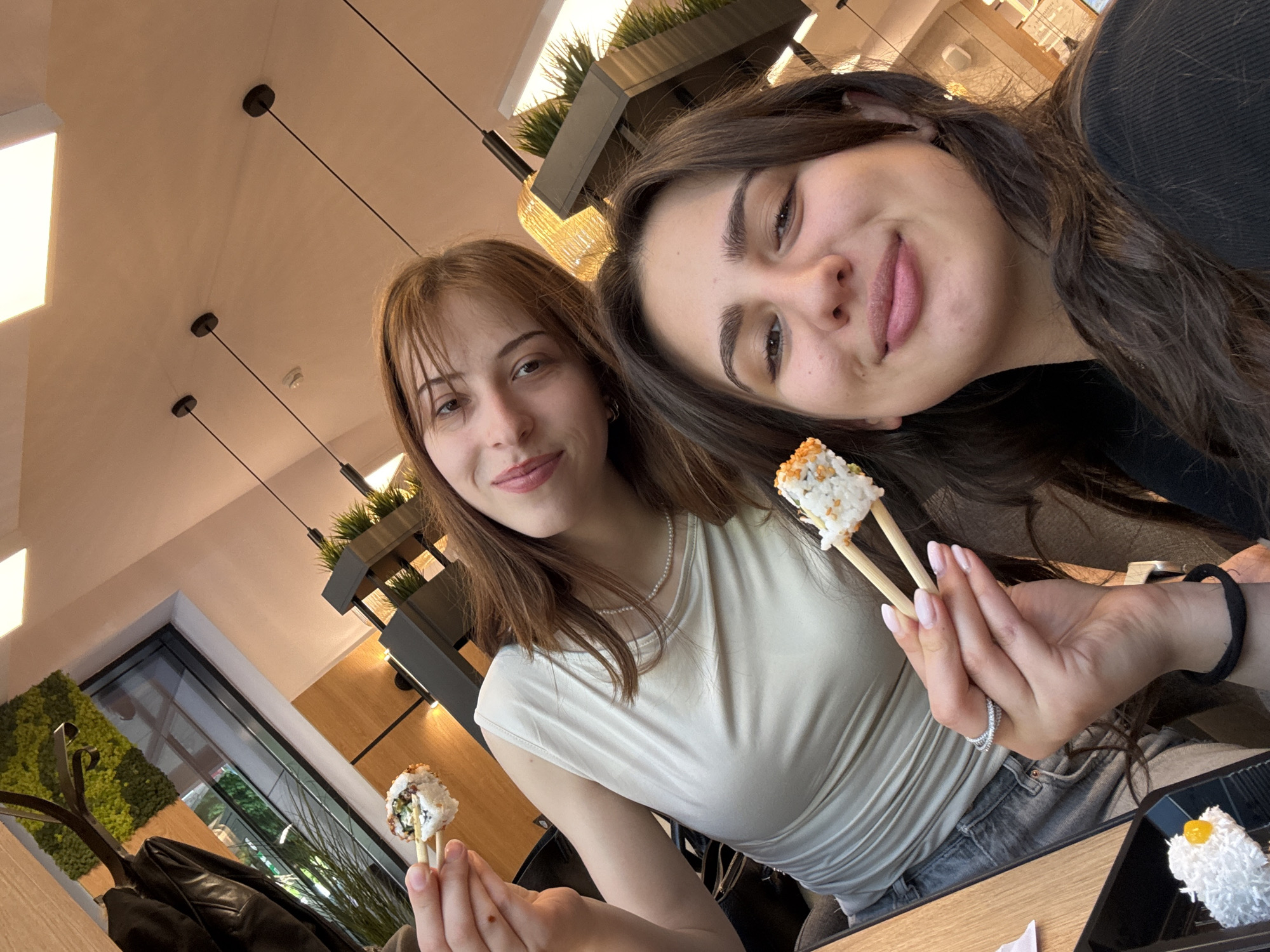 Two women are holding sushi rolls with chopsticks in an indoor setting. The woman on the left has brown hair with bangs and is wearing a light-colored top and a pearl necklace. The woman on the right has long, dark hair and is wearing a dark top. They are both smiling. A black tray with sushi is visible. The background includes hanging lights, greenery, and a window. The location is likely in Sofia, Bulgaria, based on the information provided by the user.