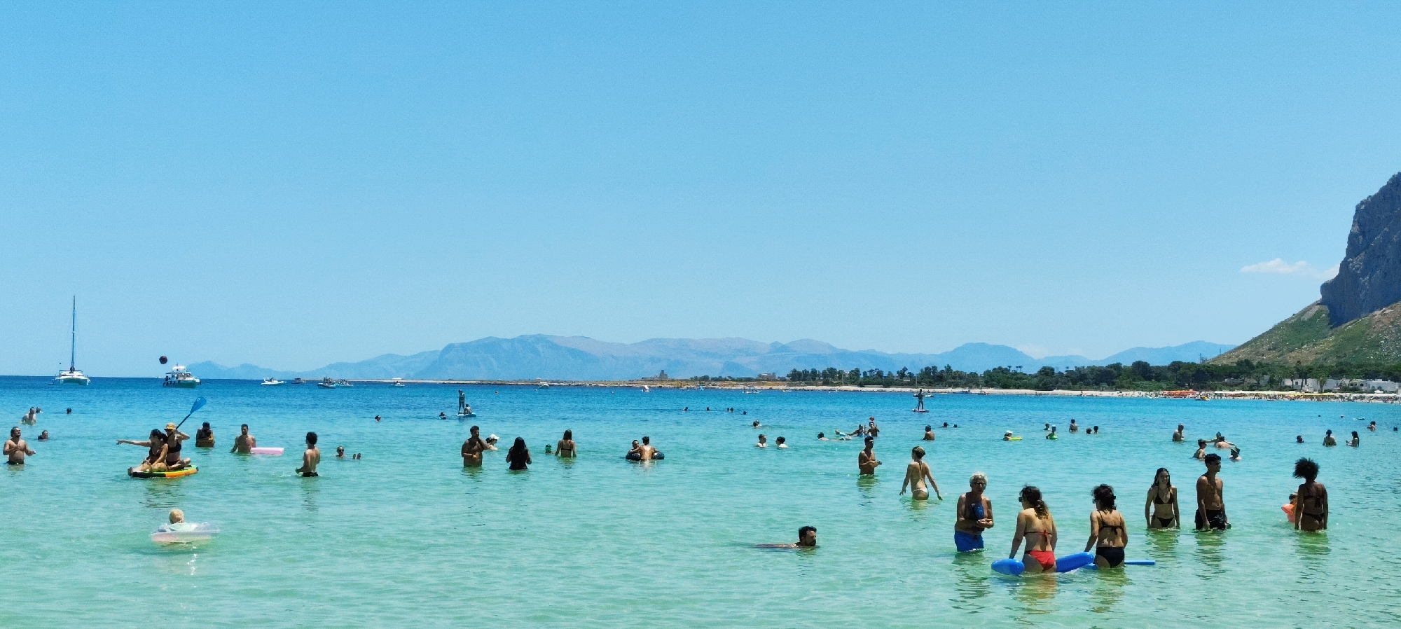 A panoramic view of a crowded beach on a sunny day, located in the city of San Vito Lo Capo, Italy. The turquoise water is clear and shallow, with a sandy bottom visible in places. Numerous people are engaged in various water activities, including swimming, wading, sunbathing on inflatable devices, and paddleboarding. A sailboat and a motorboat are visible in the background, along with a distant mountainous coastline. The sky is clear and blue, indicating good weather. The scene conveys a sense of leisure and recreation.