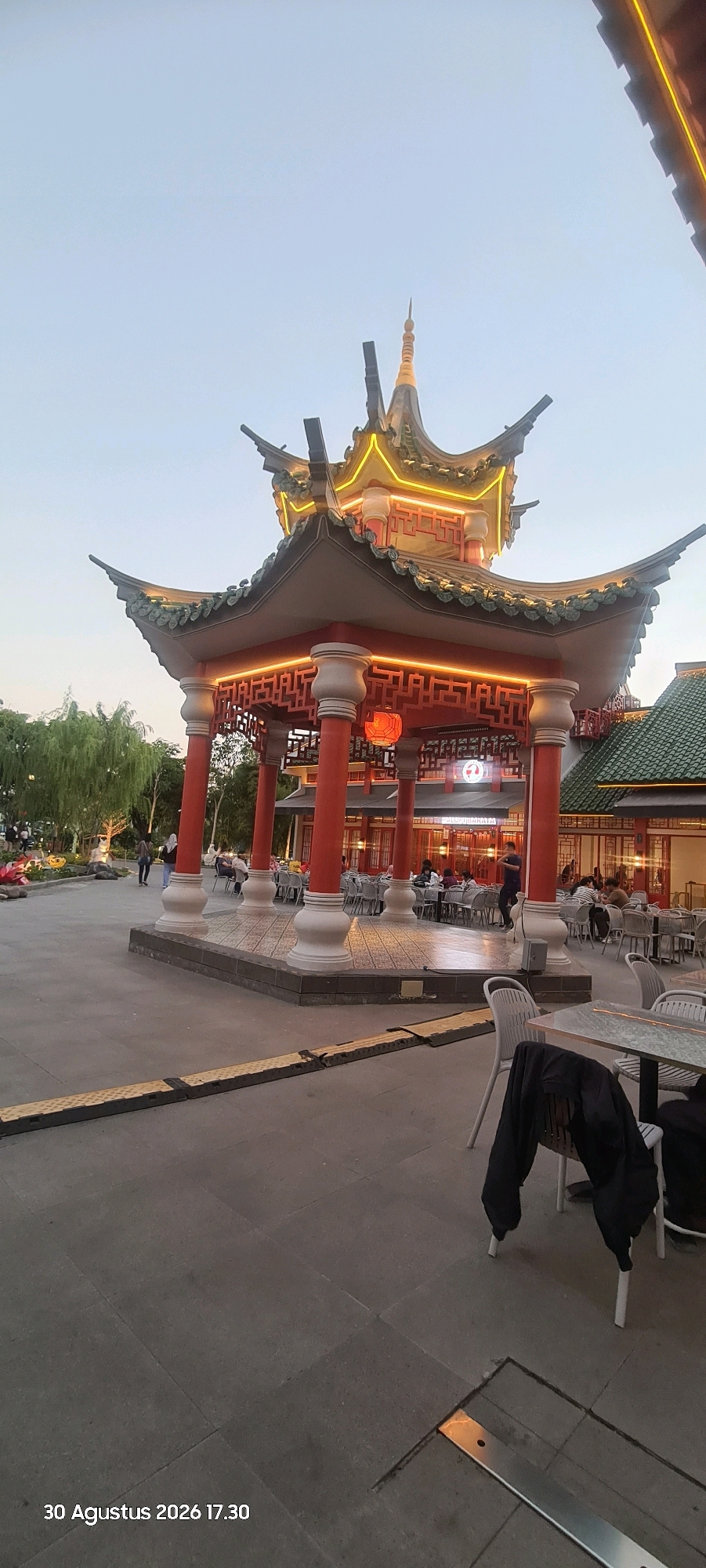 The image captures an outdoor scene in Surabaya, Indonesia, at 17:30 on August 30, 2026. The focal point is a multi-tiered, traditional Chinese-style pavilion with ornate, curved roofs adorned with green tiles and illuminated by yellow lights. The pavilion features prominent red pillars and intricate wooden latticework. In front of the pavilion, tables and chairs are set up, with several patrons seated, suggesting a restaurant or cafe environment. To the left of the pavilion, there is a landscaped area with trees and decorative elements, and some individuals are walking in the background. The ground is paved with large, dark gray tiles, and a yellow and black textured speed bump is visible in the foreground. The sky is a pale blue, indicating dusk.