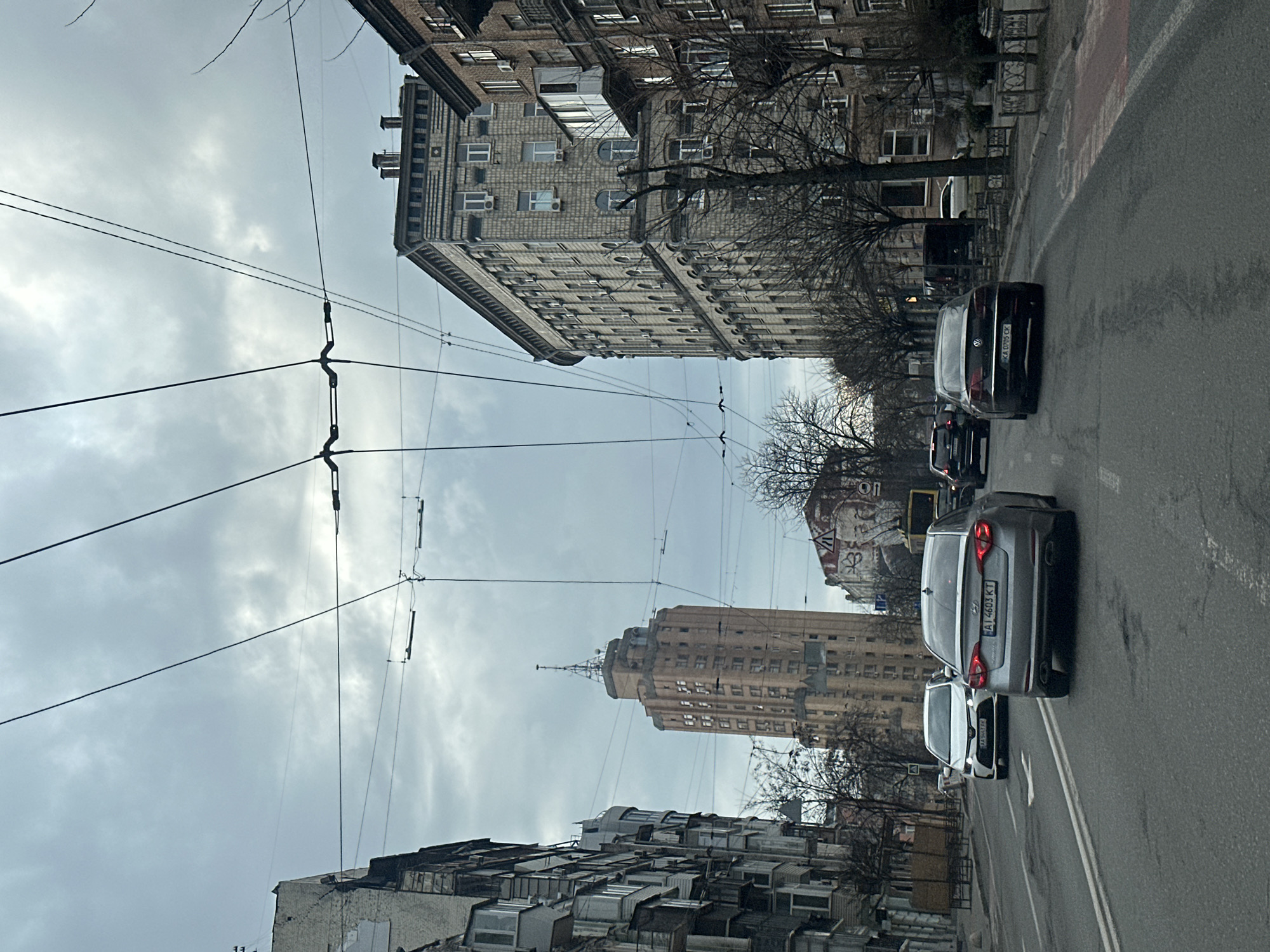 The image captures a street scene in Kyiv, Ukraine, characterized by a mix of architectural styles under an overcast sky. On either side of the road, buildings are visible, one constructed of grey brick with multiple levels of windows, and the other with older, more dilapidated features. Overhead, a web of electrical wires crisscrosses the sky.
Several cars are present on the street. There are 3 cars in focus going away from the observer. A gray Hyundai is visible, displaying the Ukrainian license plate AI 4603 KT. The license plate on a black Volkswagen can be seen and it reads KA 0705 CX. The road is marked with double white lines. A pedestrian walkway is visible to the right, bordered by trees and a stone fence.