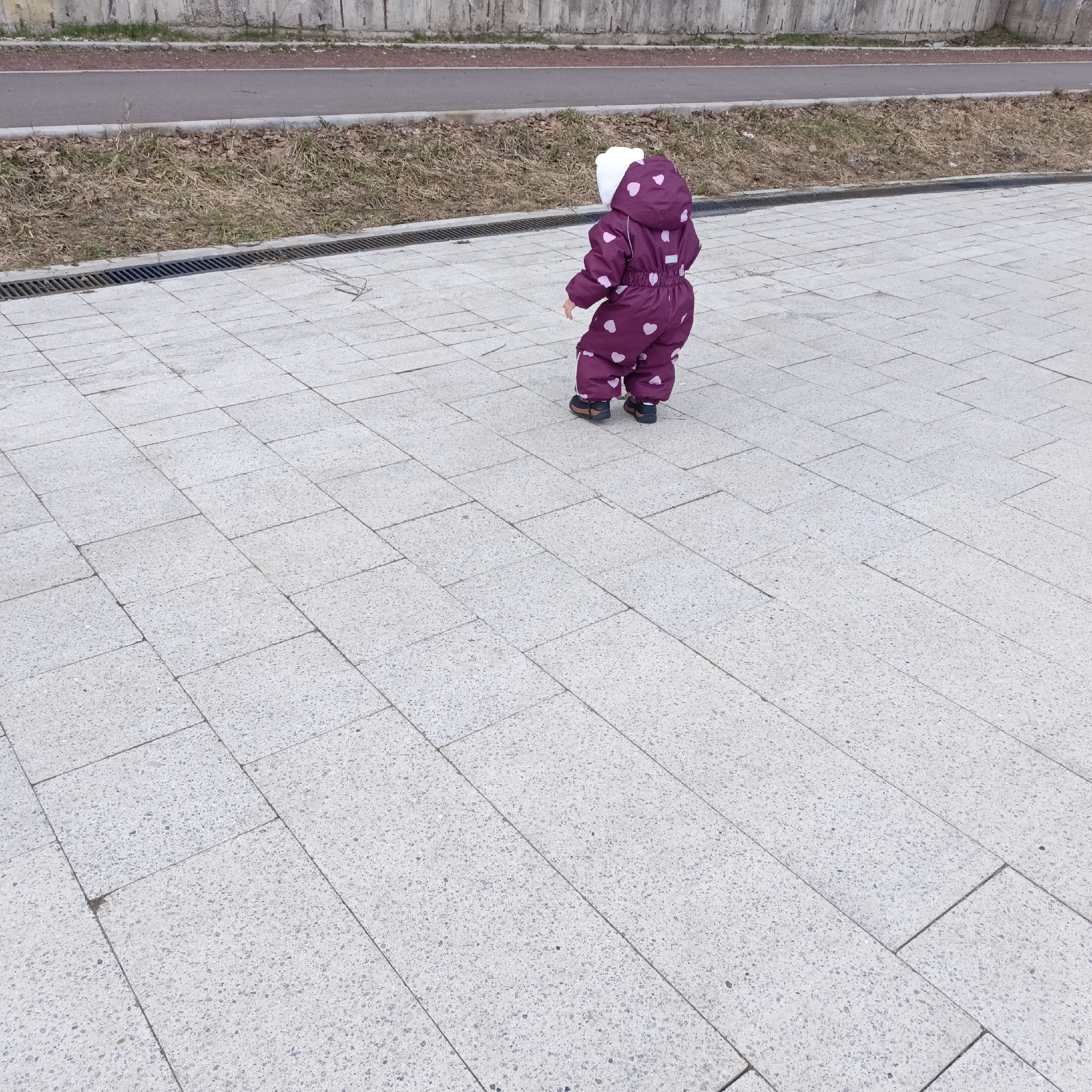 A toddler wearing a purple snowsuit with white heart patterns and a white hat stands on a light gray paved surface. A drainage grate runs parallel to a strip of dead grass and bare trees. Behind this is a narrow red path and a concrete wall. The location is Unknown, Unknown.