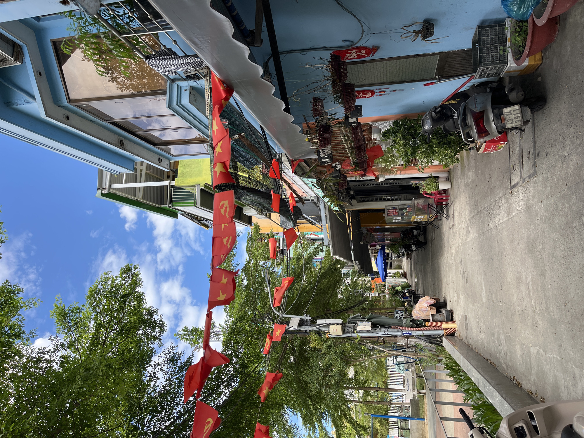 This media file captures a vibrant street scene in Đà Nẵng, Vietnam, under a bright blue sky with scattered white clouds, suggesting a sunny day. The image is taken from a low angle, looking down a narrow street or alleyway lined with multi-story buildings and lush green trees.
Numerous red flags of Vietnam with yellow stars, alongside flags of the Communist Party of Vietnam with yellow hammer and sickle, are strung across the street, creating a festive or commemorative atmosphere. The buildings feature various architectural styles, some painted a light blue, with prominent windows, balconies, and white awnings extending over storefronts. One building's large glass facade reflects the sky and trees.
The street itself is paved with concrete. Several motorcycles or scooters are parked along the side, indicating a typical mode of transport in the area. One scooter displays a license plate reading "43-XL 3911". Potted plants and other decorative items, including red "AIR" branded decorations, adorn the building fronts. Utility poles with a dense network of wires are visible, typical of Vietnamese urban infrastructure. Further down the street, a sign for "Seabank" is partially visible. The presence of shop awnings and outdoor displays suggests commercial activity, though no specific human activity or people are prominently captured. The overall scene depicts a lively, everyday street environment in an urban Vietnamese setting.