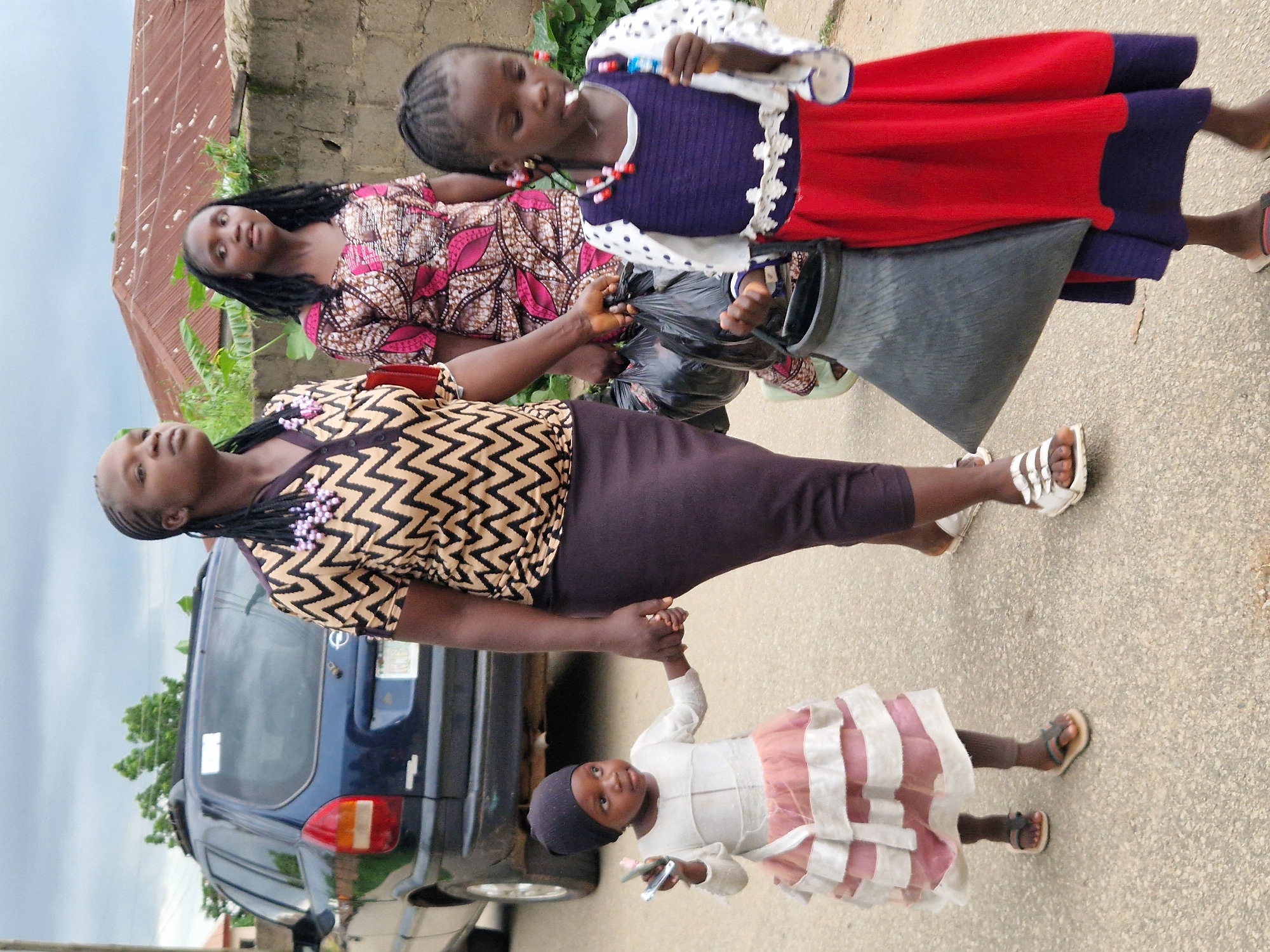 In an outdoor, urban setting with overcast skies, a group of four Black females walks on a paved surface. A dark blue SUV is parked on the left side of the frame. In the foreground, a young child in a light-colored, tiered dress and black beanie holds the hand of an adult female. This adult is wearing a patterned top with a zigzag design and a dark skirt. To the right, another adult female is dressed in a pink and white patterned dress. A young girl in a red and blue dress walks to the far right, holding a lollipop. One adult female carries two black plastic bags and a large black conical bag. The background features a brick wall and a building with a corrugated metal roof.