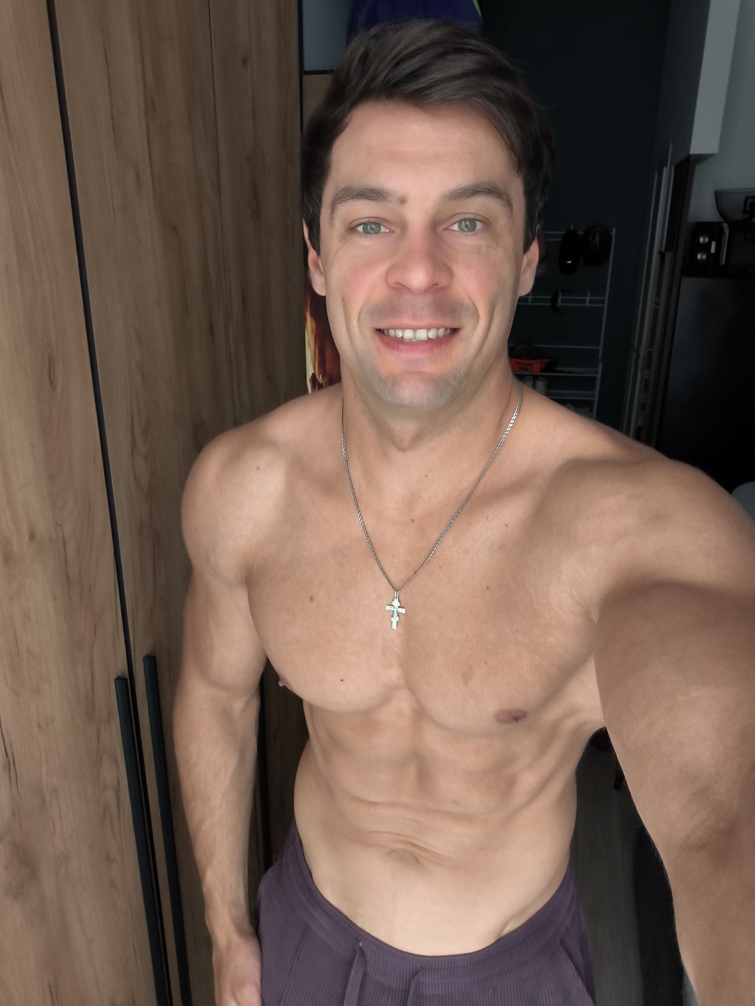 The image is a selfie taken indoors, likely in a bedroom or dressing area, with a wardrobe visible in the background. The subject is a shirtless man with visible abdominal muscles and a prominent cross necklace. He has fair skin, dark hair, and green eyes, and is smiling broadly at the camera. The lighting suggests it is daytime, though the specific time is not evident. The man is wearing dark purple sweatpants. The background includes a wooden wardrobe with dark handles and a glimpse of shelves with some objects on them in the distance. The overall impression is of a casual, self-assured individual.