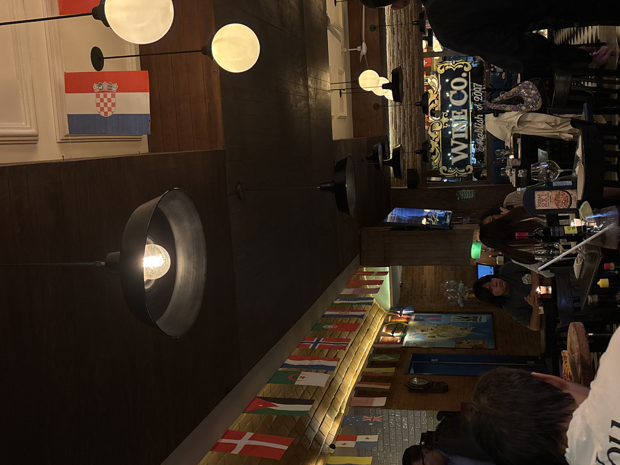 The image shows the interior of a restaurant or bar, potentially "Wine Co." established in 2007. Flags from various countries decorate the wall. A bar area is visible with a person working behind it. Customers are present, some seated at tables. The lighting is a mix of ambient light and prominent hanging light fixtures. The location is likely in Mexico City, Mexico, based on the description.