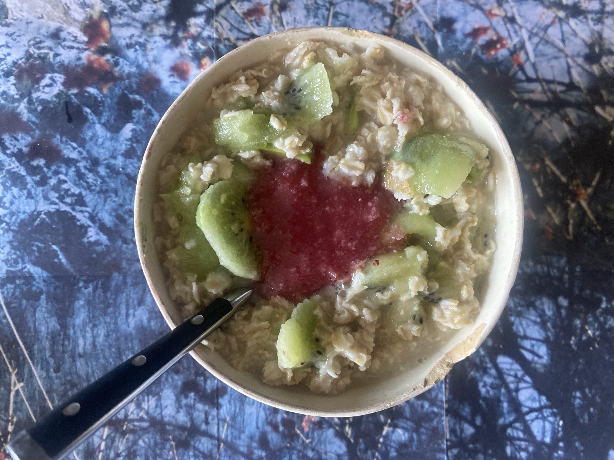 An overhead view captures a single serving of oatmeal in an off-white bowl. The oatmeal is garnished with multiple irregularly cut green kiwi slices, some displaying visible black seeds, and a central dollop of red, textured fruit puree. A silver-colored spoon with a black handle, featuring three visible rivets, rests partially submerged in the oatmeal on the left side of the bowl. The bowl exhibits a slightly rustic rim with a minor chip on its lower right edge. The bowl and its contents are situated on a patterned surface depicting a stylized winter forest scene, characterized by dark blue and white snow-like textures, dark bare tree branches, and interspersed reddish-orange elements. No individuals are visible. The scene is located in an indoor setting in Unknown, Unknown.