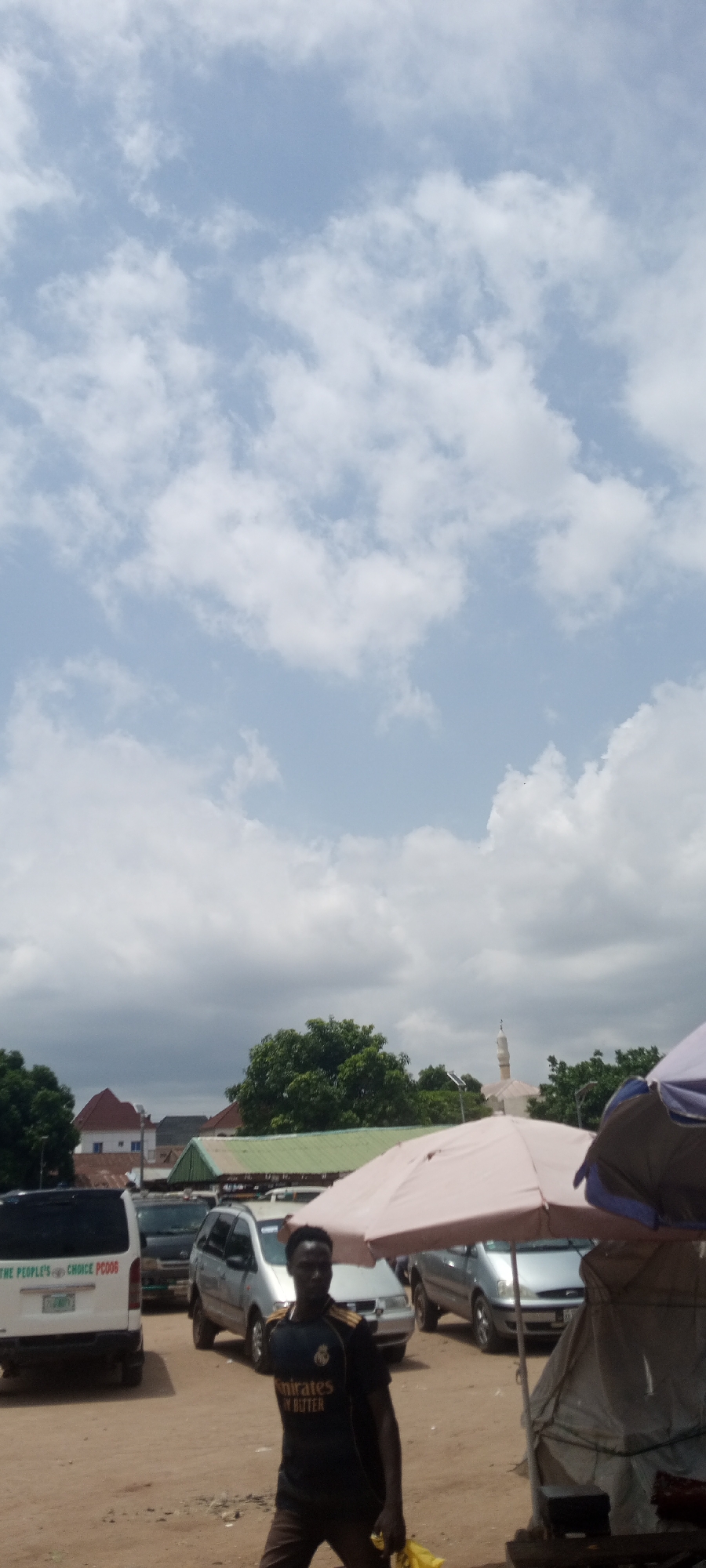 The image captures a street scene in Jalingo, Nigeria, under a partly cloudy sky, suggesting it is daytime. In the foreground, a man wearing a dark t-shirt with "Emirates FLY BETTER" text and dark pants is walking. He is holding a yellow bag in his right hand. Behind him, several vehicles are parked, including a white van with the text "THE PEOPLE'S CHOICE PC006" on its rear. Other vehicles visible are silver SUVs and a darker colored SUV. A large, light-colored umbrella is prominently displayed, providing shade. In the background, various buildings are visible, including structures with red and green roofs, and a mosque with a minaret. Lush green trees are scattered throughout the scene. The overall impression is of a bustling local area, possibly a market or a street side gathering.