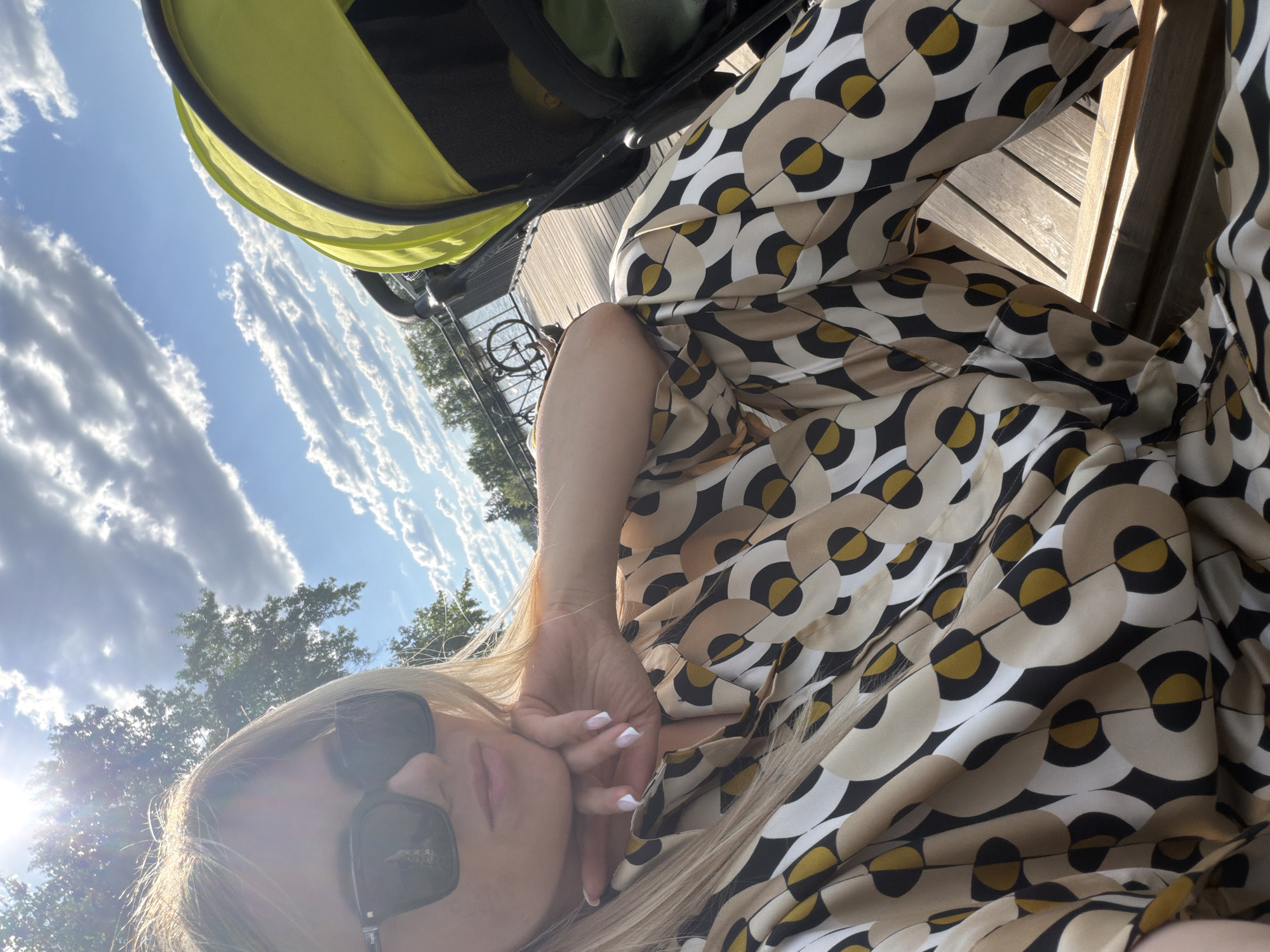 A blonde woman, wearing dark rectangular sunglasses and a long-sleeved garment with a geometric pattern of beige, white, black, and mustard yellow circles and semi-circles, is positioned in the left foreground. Her chin rests on her left hand, which exhibits a white-tipped manicure. To her right, a bright neon green and black baby stroller is partially visible. Both subjects are situated on a light brown wooden deck or promenade. A black metal railing separates the deck from a body of water in the mid-ground. A bicycle is secured to the railing near the water's edge. Trees line the distant horizon across the water. The sky is clear blue with scattered white cumulus clouds, particularly dense on the right. A strong lens flare from the sun is present in the upper left quadrant, indicating bright daylight conditions. The scene is an outdoor setting in Votkinsk, Russia.