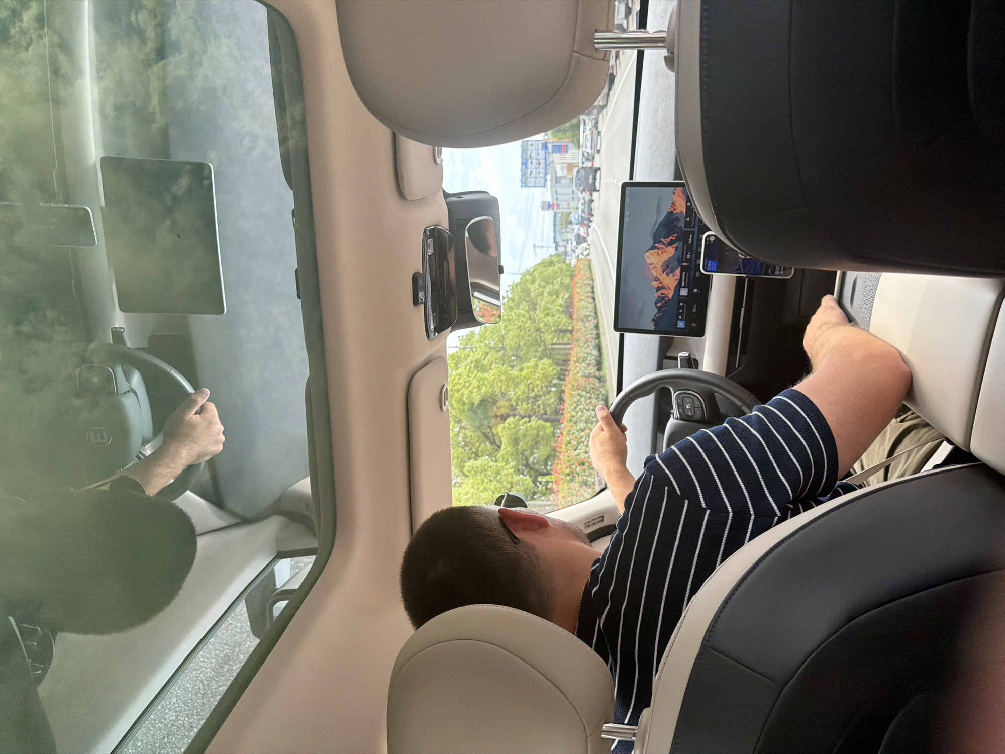 The image shows the interior of a car driving on a road, with the driver visible from the back. The driver is a man with short dark hair, wearing sunglasses and a navy and white striped shirt. He is holding the steering wheel, and his right arm is resting on the center console. The car has a panoramic sunroof, through which the sky and surrounding trees are visible. In the foreground, the back of the driver's seat and the passenger seat are visible. On the dashboard, there is a large touchscreen display showing a mountain landscape image, and a smartphone mounted to the dashboard. The car is in traffic, with other vehicles visible on the road ahead. The environment appears to be a city street with trees and buildings lining the road. Based on the presence of billboards with Russian text, the location is likely Sochi, Russia.