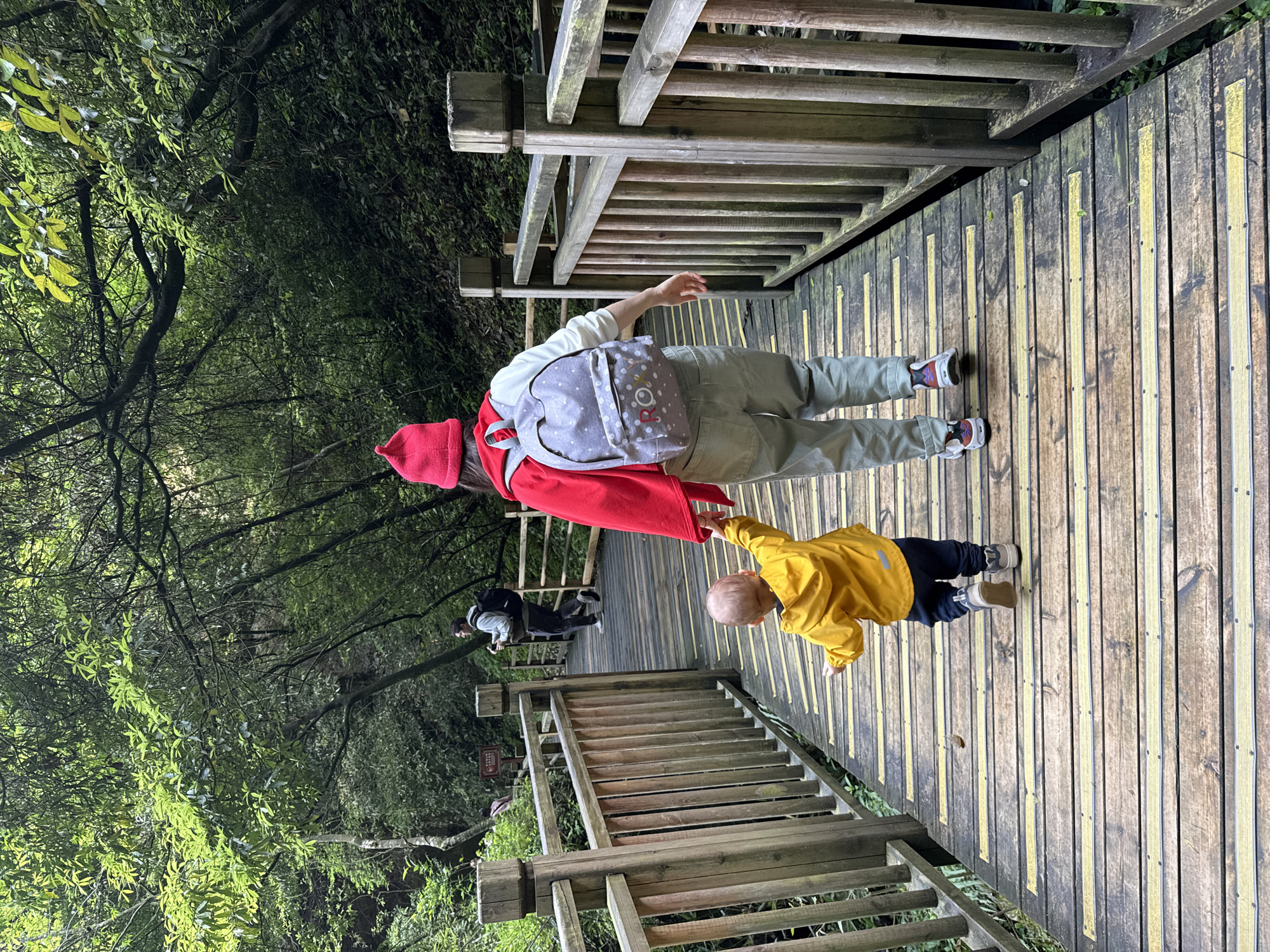 On a wooden boardwalk in the city of Zhangjiajie, China, a person wearing a red hat, red and white sweater, grey pants, and a grey backpack is holding the hand of a small child wearing a yellow jacket and blue pants. The boardwalk is surrounded by a dense forest. Another person, dressed in darker clothing, is further down the boardwalk in the same direction. The boardwalk has wooden railings on either side.