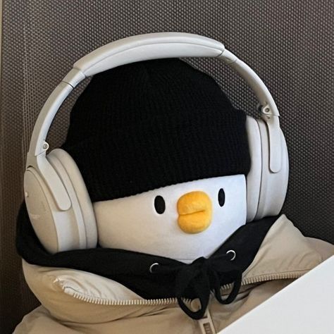 The image shows a plush toy wearing a black beanie and white headphones. The plush toy's head is white with a yellow beak and two black eyes. The plush toy is wearing a brown jacket and black hoodie. The background is a gray textured surface. The image appears to have been taken indoors. The coordinates (-7.45472, 111.427584) indicate a location near the city of Geneng, Indonesia, however, the image does not provide any further details about the location. There are no people in the image.