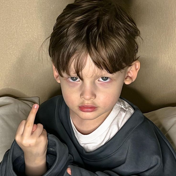 The image shows a young boy with brown hair, wearing a grey sweatshirt over a white t-shirt, giving the middle finger. The boy is sitting on a bed with a tan headboard and a white pillow. He has a serious expression on his face. The coordinates provided do not reveal any information about the location of this photo. It is not possible to determine where the photo was taken.