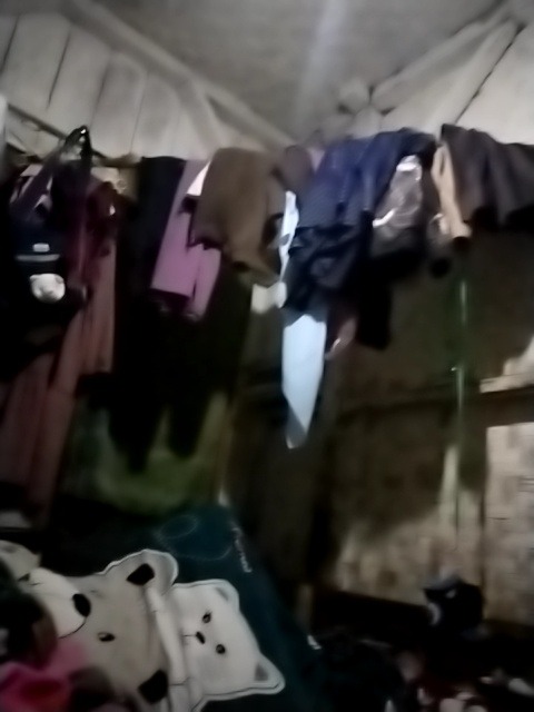 The image shows an indoor scene with clothes hanging on a clothesline. The clothes are of various colors and types. In the foreground, there is a dark colored blanket with a pattern of white teddy bears. The background is dimly lit and appears to be a wall. The scene is consistent with a residential dwelling in Campakawarna, Indonesia, likely a bedroom or living area where clothes are dried indoors. There are no people visible in the image. The overall impression is one of domesticity and everyday life in a modest setting.