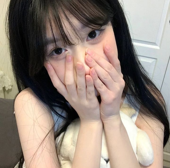 The image shows a young woman with long black hair covering most of her face. Her hands are covering her mouth and nose. She is wearing a white tank top. The image was taken indoors, possibly in a bedroom. The woman's facial expression suggests she may be feeling shy or embarrassed. The coordinates provided (-7.45472, 111.427584) correspond to a location in the city of Geneng, Indonesia.