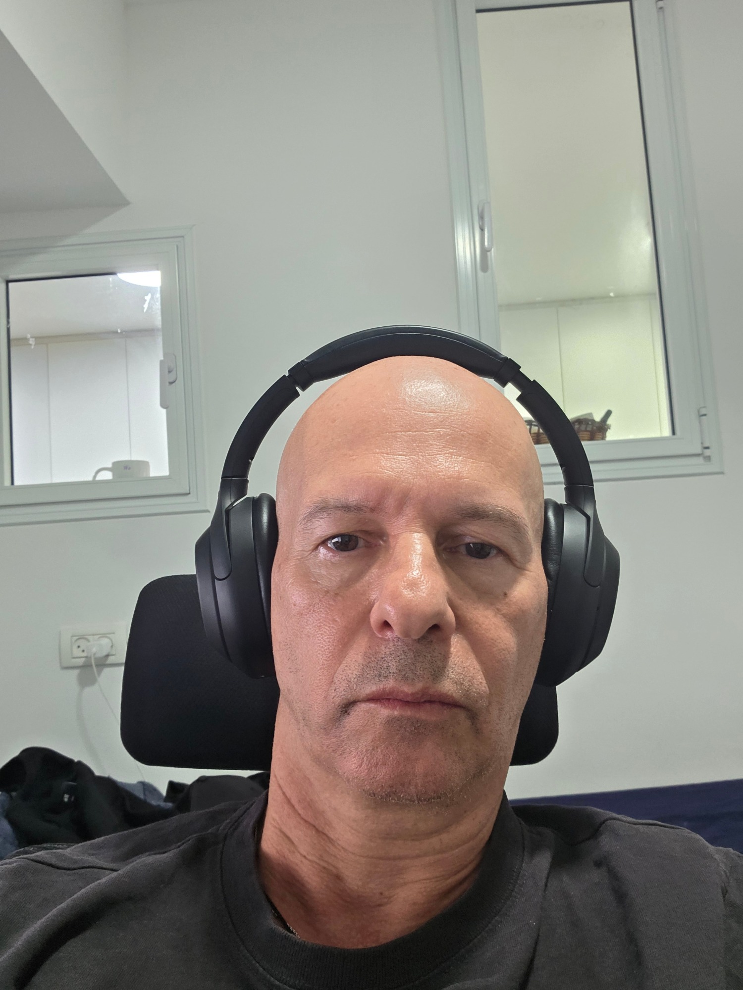 A bald, middle-aged man with headphones on is seated in an office chair. He is wearing a black t-shirt. In the background, there are windows and a wall-mounted electrical outlet. The scene appears to be indoors, likely in an office or home setting, given the furniture and fixtures. The lighting suggests it is daytime. The overall impression is a casual, personal photograph taken by the subject himself.