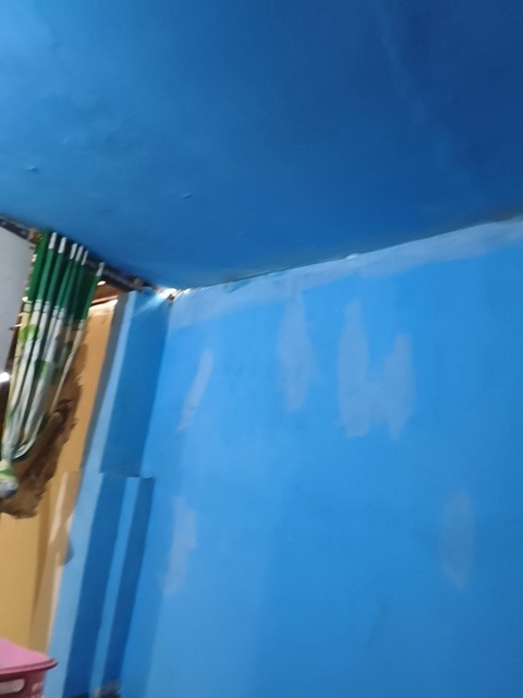 The image depicts an interior scene, likely a room, with a dominant blue color scheme on the walls and ceiling. The ceiling is a solid, bright blue, showing some minor imperfections and possibly slight texturing. The walls are also painted blue, with several irregular, lighter patches that suggest areas that may have been patched or are showing through the paint. To the left of the frame, partially visible, are vertical blinds or curtains with a green and white striped pattern. Adjacent to the blinds is a section of wall painted a muted yellow or tan color. A corner of a pink object, possibly a bin or container, is visible in the lower left of the image. The lighting suggests it might be evening or indoors with artificial light. There are no people or animals visible in the image. The scene appears to be domestic, with a focus on the painted surfaces and the blinds. The provided crops highlight different sections of the blue ceiling and walls, as well as the detail of the blinds and the adjoining yellow wall. The location in Makassar, Indonesia, is not visually discernible from the image itself.