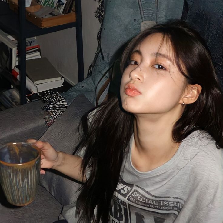 The image shows a young woman with long, dark hair sitting on a couch, holding a brown ceramic mug in her right hand. She is wearing a gray t-shirt with a black print. She is looking at the camera. The image is taken from the right side of the woman's face. The couch is covered in gray and black fabric. Behind her, a wooden bookshelf is visible, with books and a small wooden box on it. The location is a residential area in Geneng, Indonesia, based on coordinates (latitude: -7.45472, longitude: 111.427584). The lighting is dim and the background is out of focus.