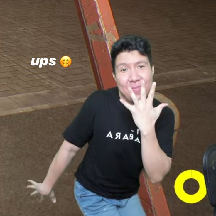 A young man wearing a black t-shirt with white lettering that says "AYA BA" and blue jeans is standing with his left hand up, his fingers spread and his right hand pointing toward the camera. He is standing next to what appears to be a large piece of wood, possibly a part of a structure or piece of furniture. The background is a blurry image of a tiled floor. The image is a still from a video, and the word "ups" with a yellow smiley face is visible on the left side of the image. The image is taken in the city of Geneng, Indonesia, at the coordinates -7.45472 latitude and 111.427584 longitude. The man is looking directly at the camera. His expression is one of surprise or alarm, and the word "ups" suggests that something unexpected has just occurred.