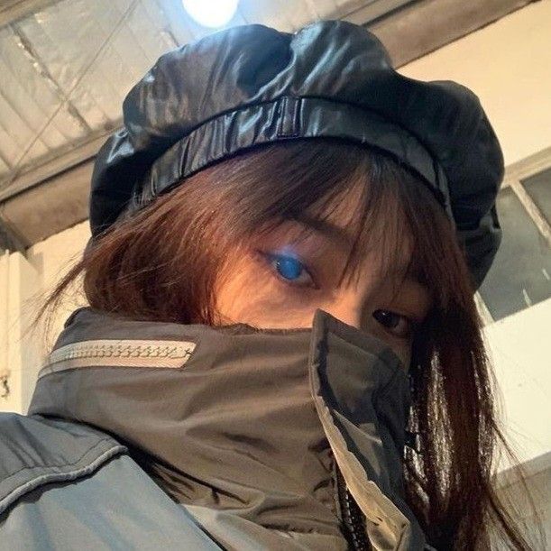 A young woman with long brown hair is wearing a black beret and a dark jacket with a zipper detail. Her face is mostly obscured by the jacket's hood, but one eye is visible, staring directly at the camera. The background is out of focus, but appears to be a building with a window. The photo was taken at a location in the city of Geneng, Indonesia, at coordinates -7.45472, 111.427584.