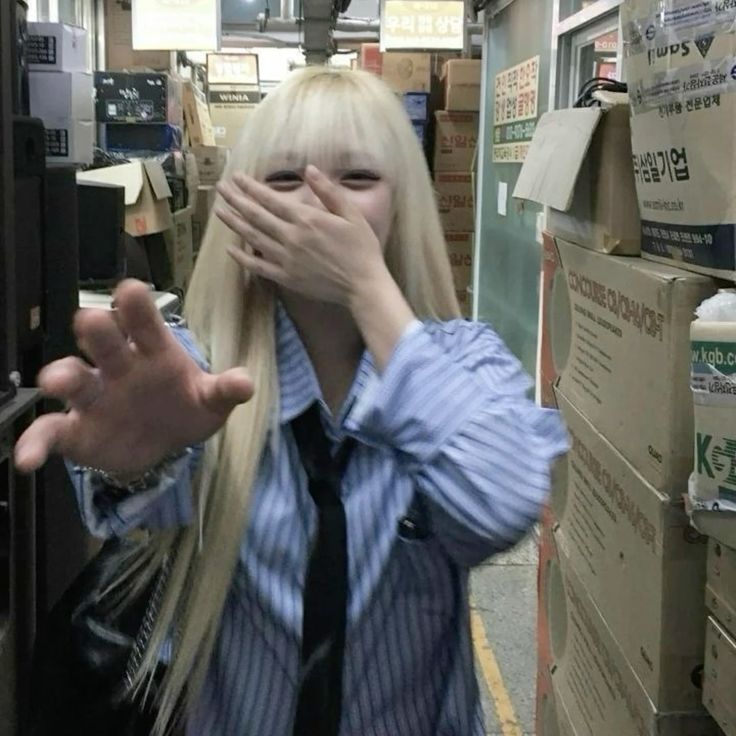 A young woman with long blonde hair and bangs is standing in a narrow corridor of a shop in Geneng, Indonesia. The woman is wearing a blue striped shirt with a black tie and is covering her face with her right hand. Her left hand is extended towards the viewer. The corridor is lined with stacks of cardboard boxes. The woman's expression is obscured by her hand, but she appears to be smiling or laughing. The shop is located at the coordinates -7.45472, 111.427584.