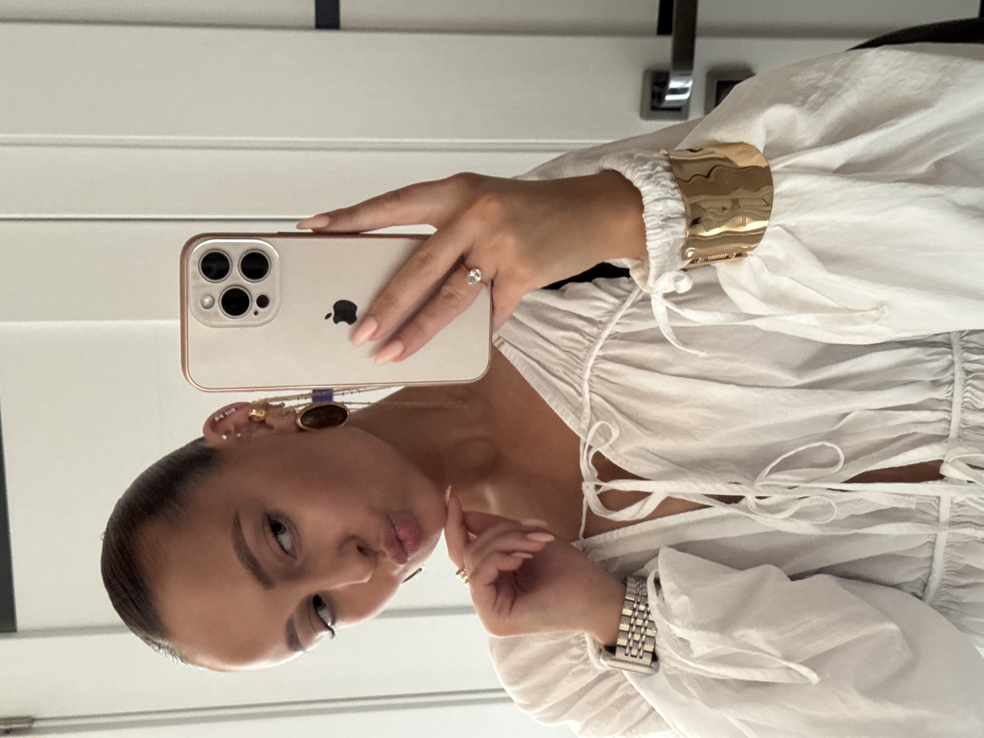 A woman is taking a selfie in a mirror. She is wearing a white, loose-fitting blouse with dramatic puffed sleeves and a tie-front neckline. She has her hair pulled back tightly and is wearing large, dangling earrings and a gold cuff bracelet. She is also wearing a silver watch and a ring on her right hand. The background appears to be an interior setting with white doors and doorframes, suggesting a domestic or hotel environment. The lighting is bright and natural.