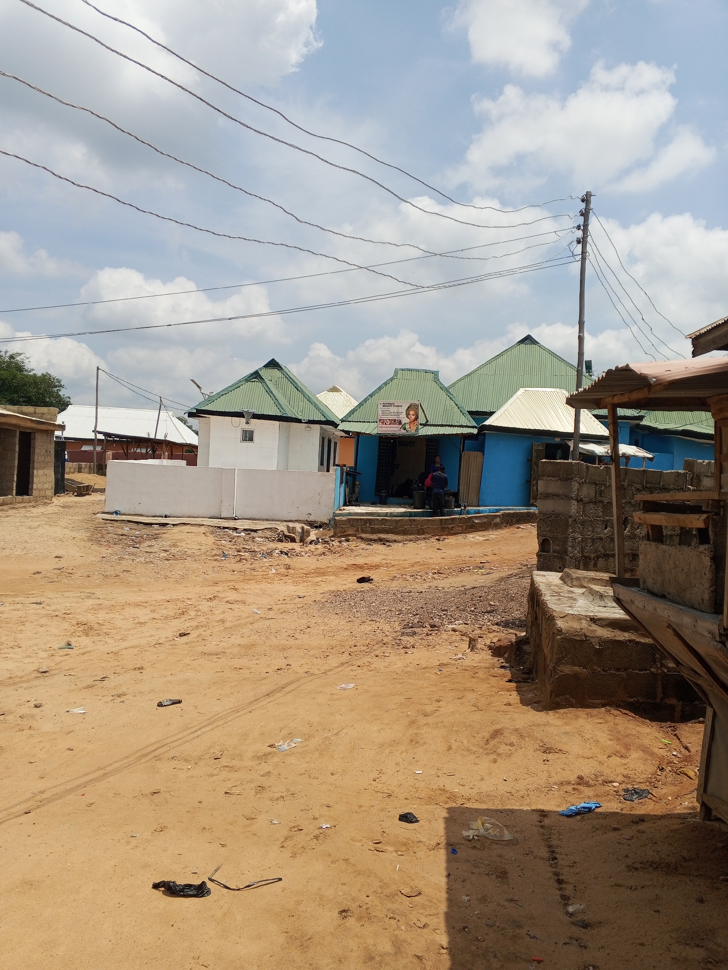 The image depicts a street scene in what appears to be a low-income neighborhood in Lokoja, Nigeria. The ground is sandy and littered with trash. Several small buildings are visible, some painted blue and white with green metal roofs. One building has a sign indicating a beauty studio. Power lines run overhead. There are indications of human activity, including people near the beauty studio. A partially constructed or decaying structure is visible in the foreground on the right.