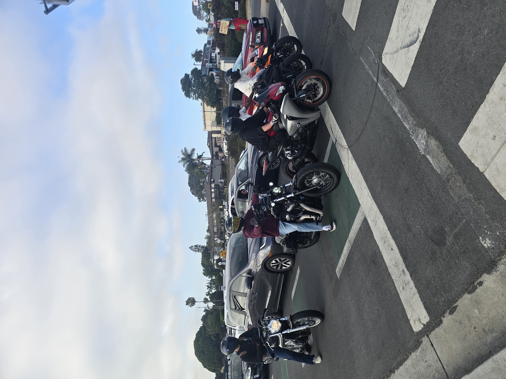 The image depicts a group of motorcyclists stopped at a crosswalk, seemingly waiting for traffic to clear or a signal to change. Four motorcycles are visible in the foreground, each with a rider wearing a helmet. The motorcycles vary in style and color, with one being a lighter metallic silver, another black, and two more in darker shades. Behind them, several cars are also stopped, indicating a traffic situation.
In the background, a person is holding a sign that reads "HOMELESS VETERAN." The environment appears to be an urban or suburban setting, with buildings, trees, and streetlights visible. The sky is partly cloudy, with a mix of white clouds and blue sky. The road surface is asphalt with clear white and green lane markings, suggesting a well-maintained street. The overall scene suggests a moment of pause in traffic, with various vehicles and individuals engaged in their own activities, including a person drawing attention to a social issue.
While the precise location within Encinitas is not explicitly identifiable from the image alone, the presence of palm trees and the general architecture could be indicative of Southern California. The street signage, if legible, would be crucial for precise identification.