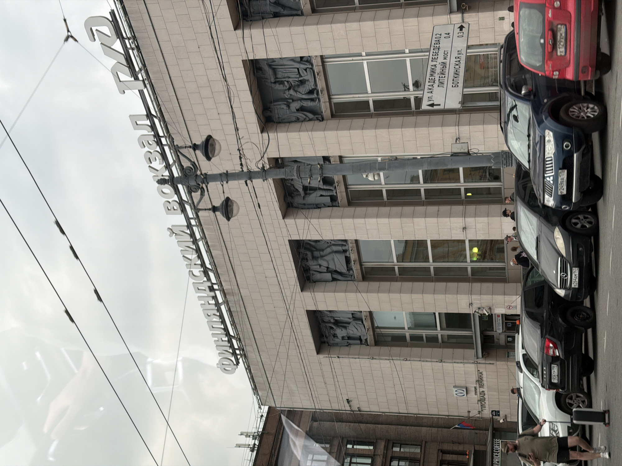 The image shows the Finland Station in Saint Petersburg, Russia, under a cloudy sky. The station's facade is adorned with a large sign reading "ФИНЛЯНДСКИЙ ВОКЗАЛ" and the RZD logo. Relief sculptures are visible above the large windows. A street light with two lamps stands in front of the building. Several cars are parked on the street, including a blue Mitsubishi SUV, a black Lada sedan, and a white SUV. A man with a backpack and a suitcase is walking on the sidewalk in front of the cars. A traffic sign with directional information is also visible. The overall scene depicts a busy urban environment with a focus on the historical architecture of the train station.