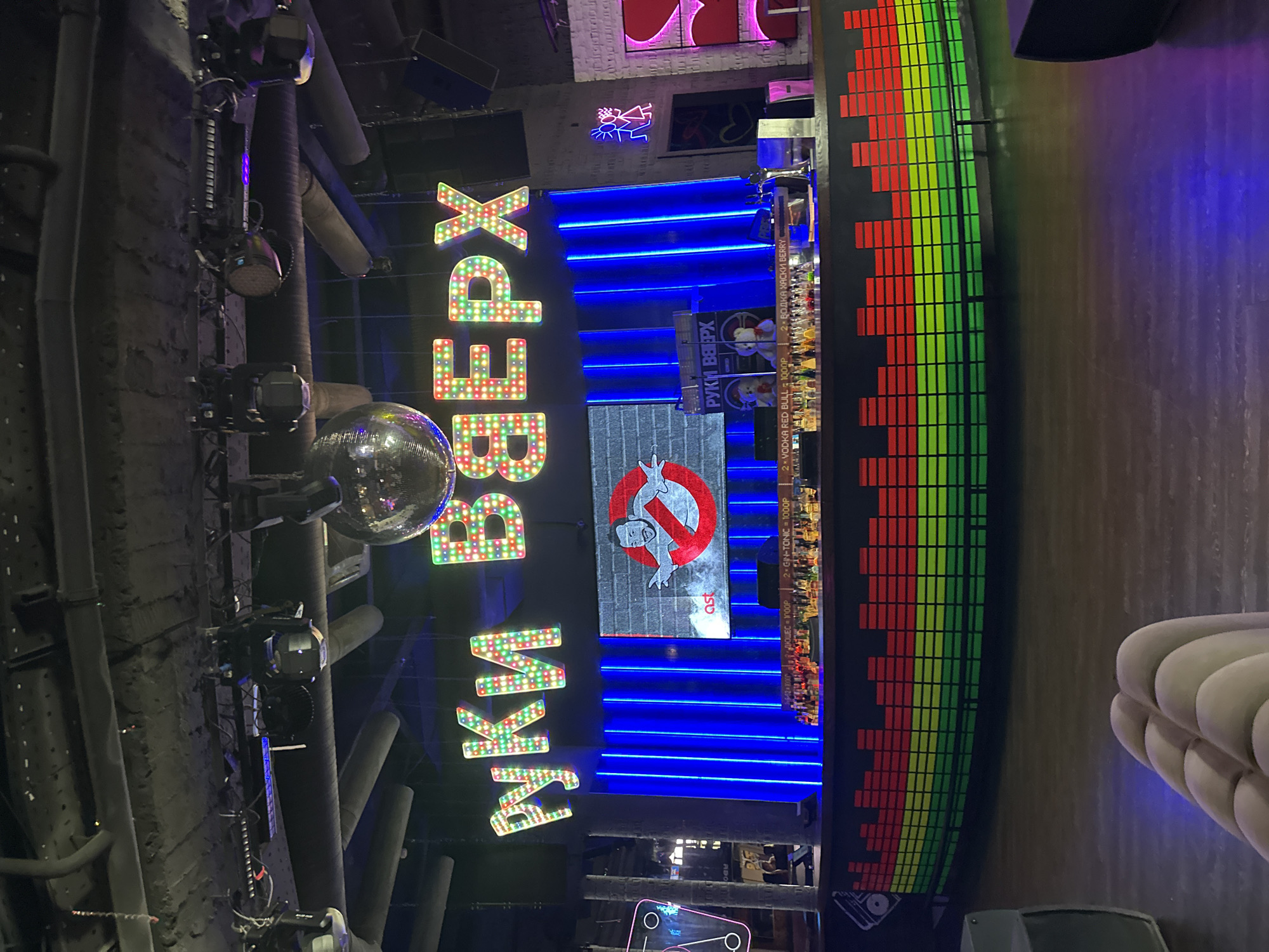 The image captures the interior of a bar or nightclub in Moscow, Russia. The name of the establishment, "Руки ВДЕРХ" (Ruki Vverkh), is prominently displayed in large, illuminated letters. A disco ball hangs from the ceiling near lighting fixtures. An image of the Ghostbusters logo is placed on a wall section. The background includes blue neon lights and a colorful audio equalizer display. A shelf stocked with bottles, likely a bar, is visible. Text in Russian indicates drink options such as "Vodka Red Bull" and "Gin-Tonic" with associated prices.