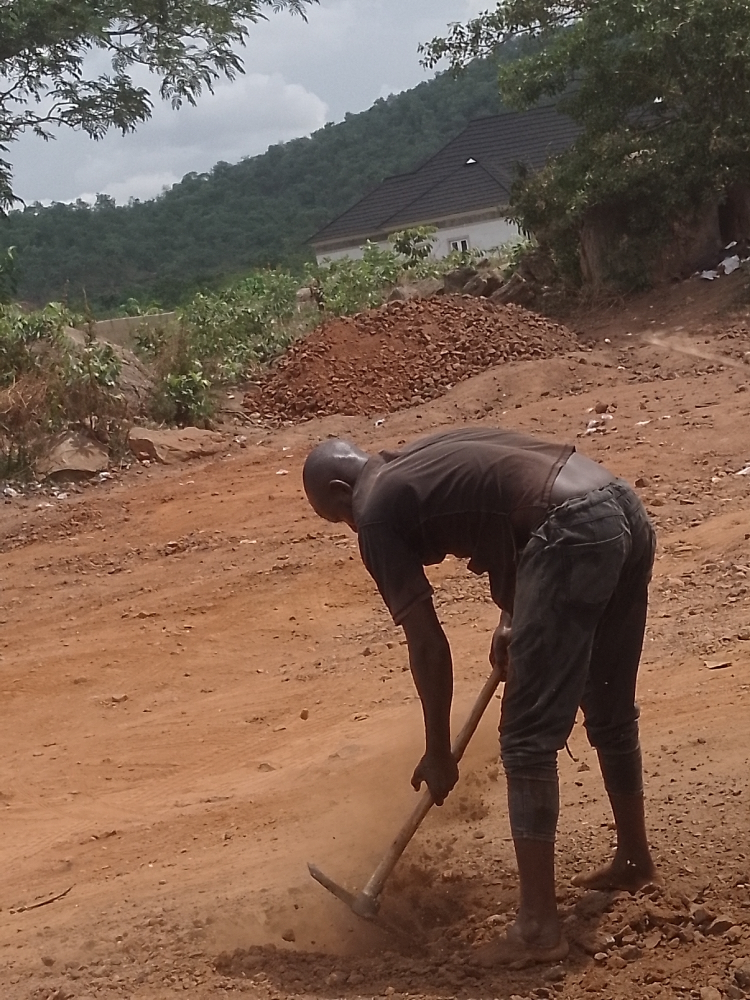 A person is working on a dirt road in Lokoja, Nigeria. The person is bald and wearing a dark-colored t-shirt and dark pants. They are using a pickaxe to break up the ground. In the background, there is a pile of red dirt and a house with a dark roof. Beyond that, a tree-covered hill can be seen. The sky is overcast.
