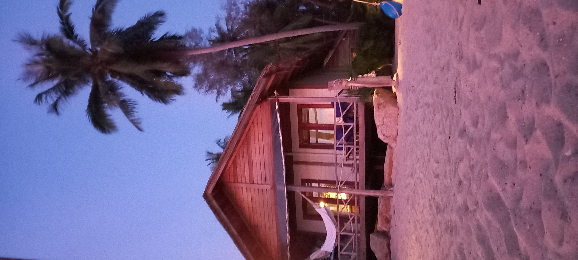 The image depicts a stilted wooden bungalow on a beach in Baan Sri Thanu, Thailand, likely at dusk. The bungalow has a gabled roof, wooden siding, and a small porch with railing and a hammock. The bungalow is illuminated with warm interior lights visible through its windows. A large palm tree is visible to the left, with its trunk extending diagonally across the upper portion of the image. The foreground consists of sandy beach.