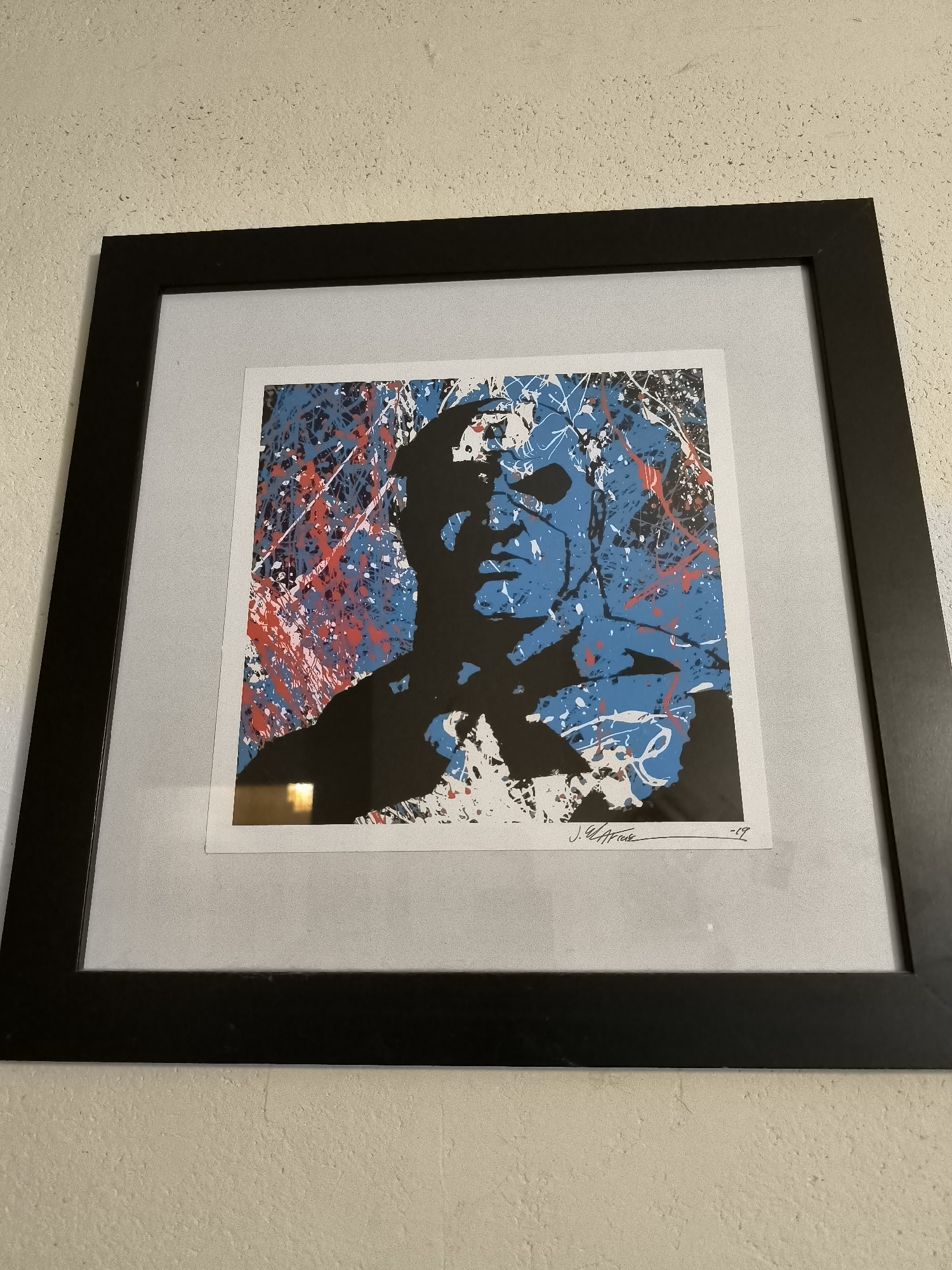 A framed artwork hangs on a beige wall. The artwork, under glass, features a portrait in black silhouette against a splatter-paint background of blue, white, and red. The portrait appears to be a human face. Below the portrait are signatures or text, partially obscured by the frame. The frame is matte black and rectangular, with a thick border. The environment appears to be an interior space, likely a home or gallery. There is no other observable details about the location in Burgas, Bulgaria, or other contextual cues.